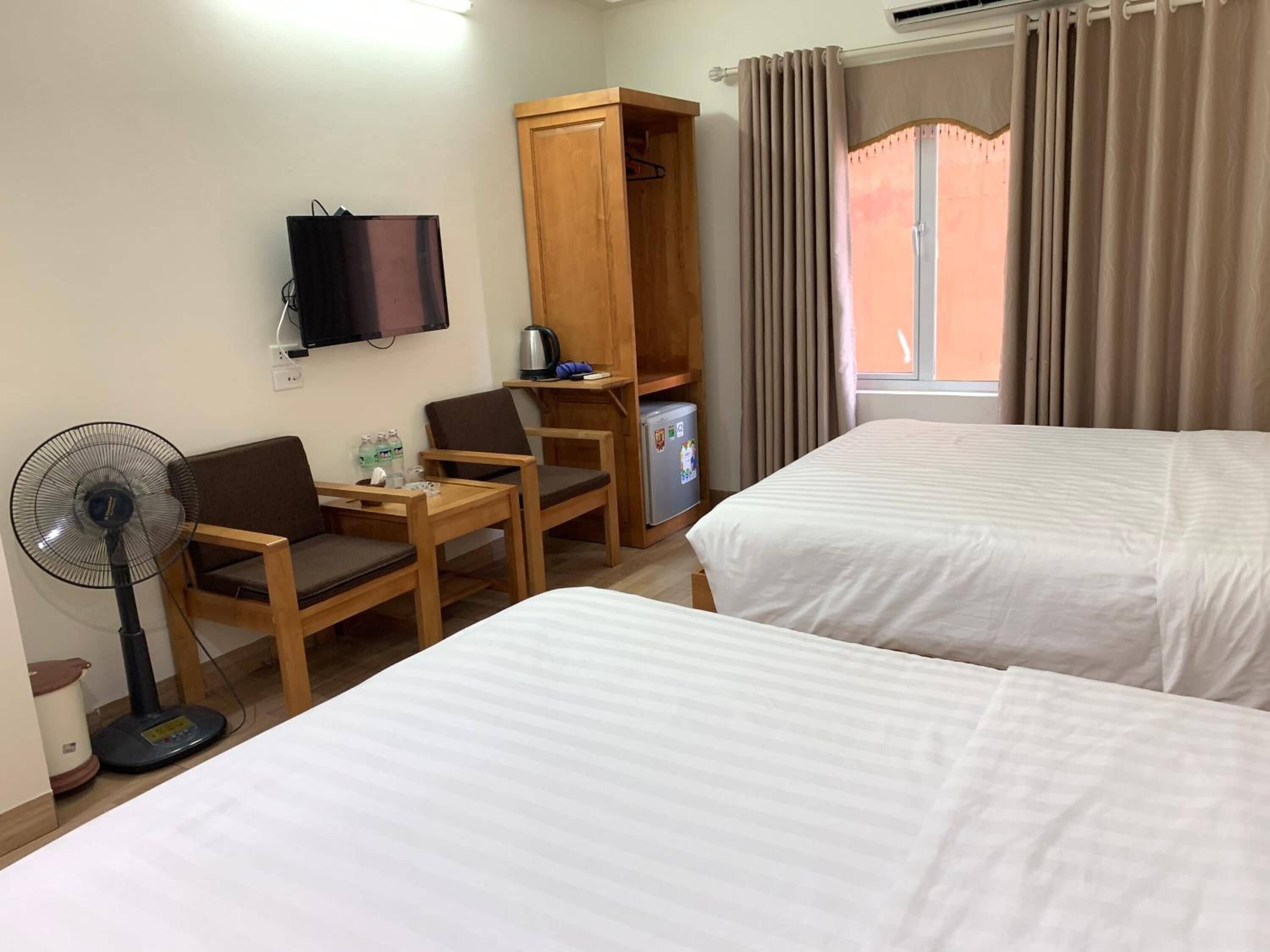 Ein Hotelzimmer mit zwei Betten, einem Schreibtisch mit Stuhl, einem Fernseher, einem Kleiderschrank, einem Minikühlschrank und einem Ventilator.