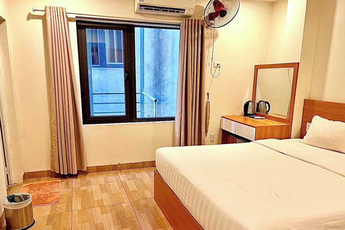 A hotel room with a bed, a bedside table with a lamp, a window with curtains, a fan, and a trash bin.