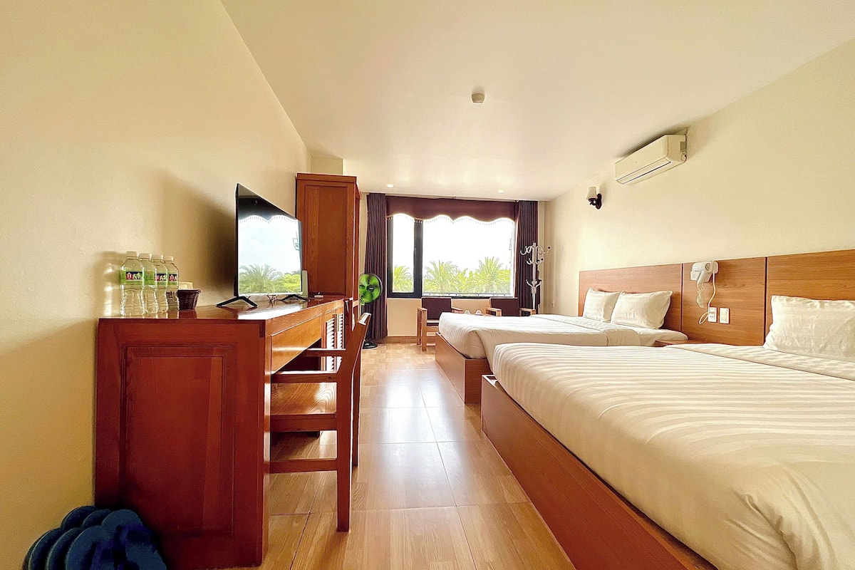 A hotel room with a wooden desk, two beds, a TV, and a window with a view of greenery.