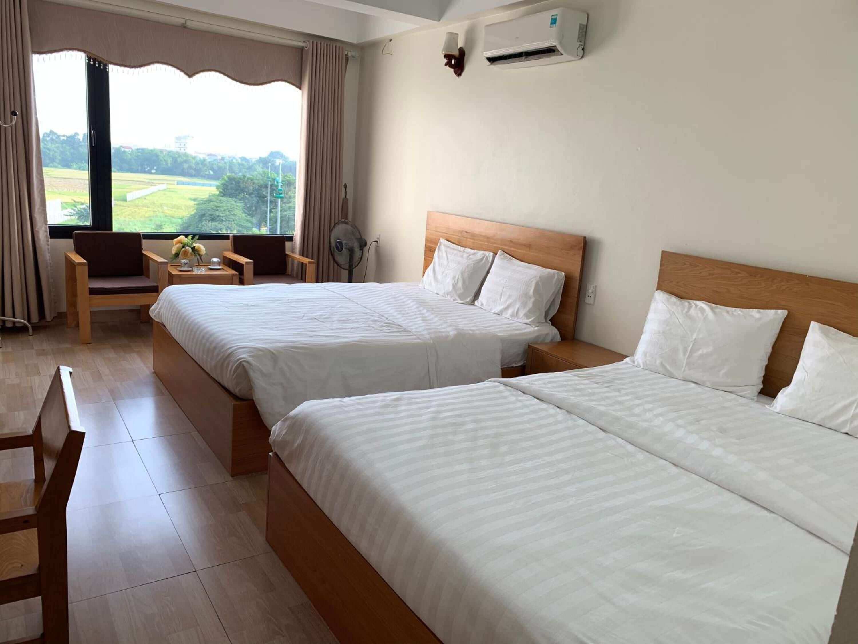 A hotel room with two beds, a desk, a chair, an air conditioner, and a view of a green landscape.