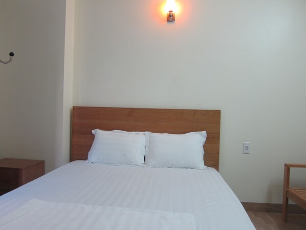 Una cama bien hecha con sábanas blancas, cabecera de madera y una lámpara fijada a la pared.