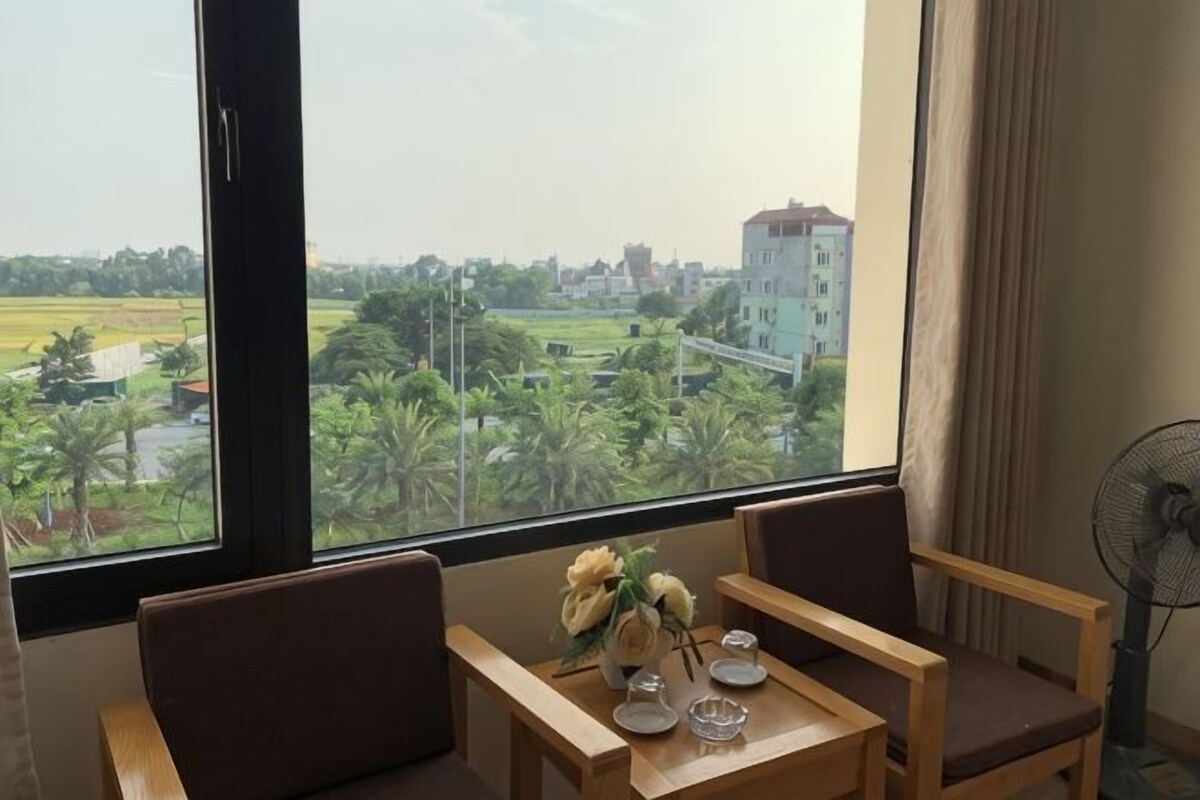 Una habitación con un gran ventanal, una mesa con sillas, un florero con flores y un ventilador.