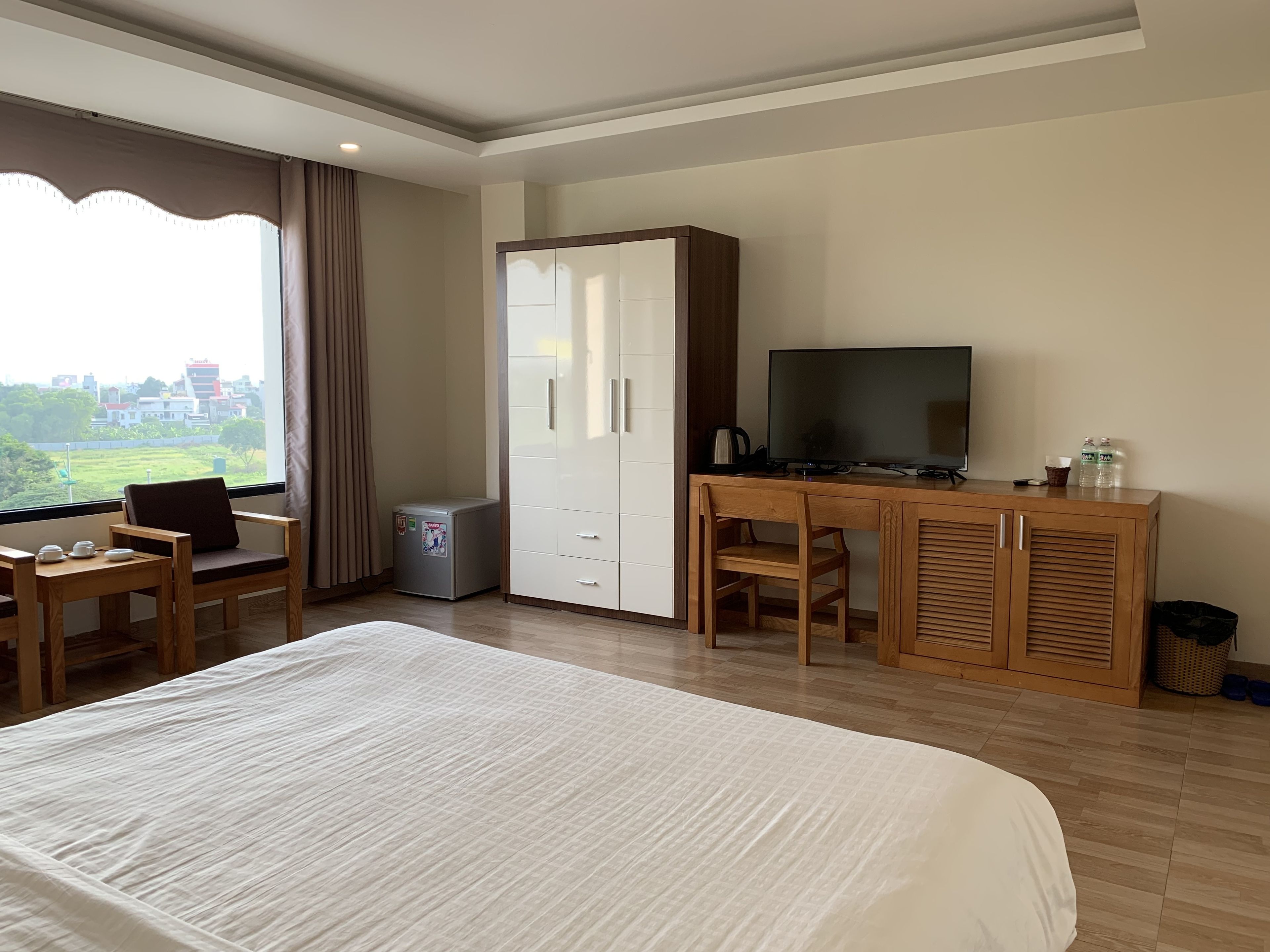A hotel room with a large bed, a desk with a television, a wardrobe, and a small refrigerator.