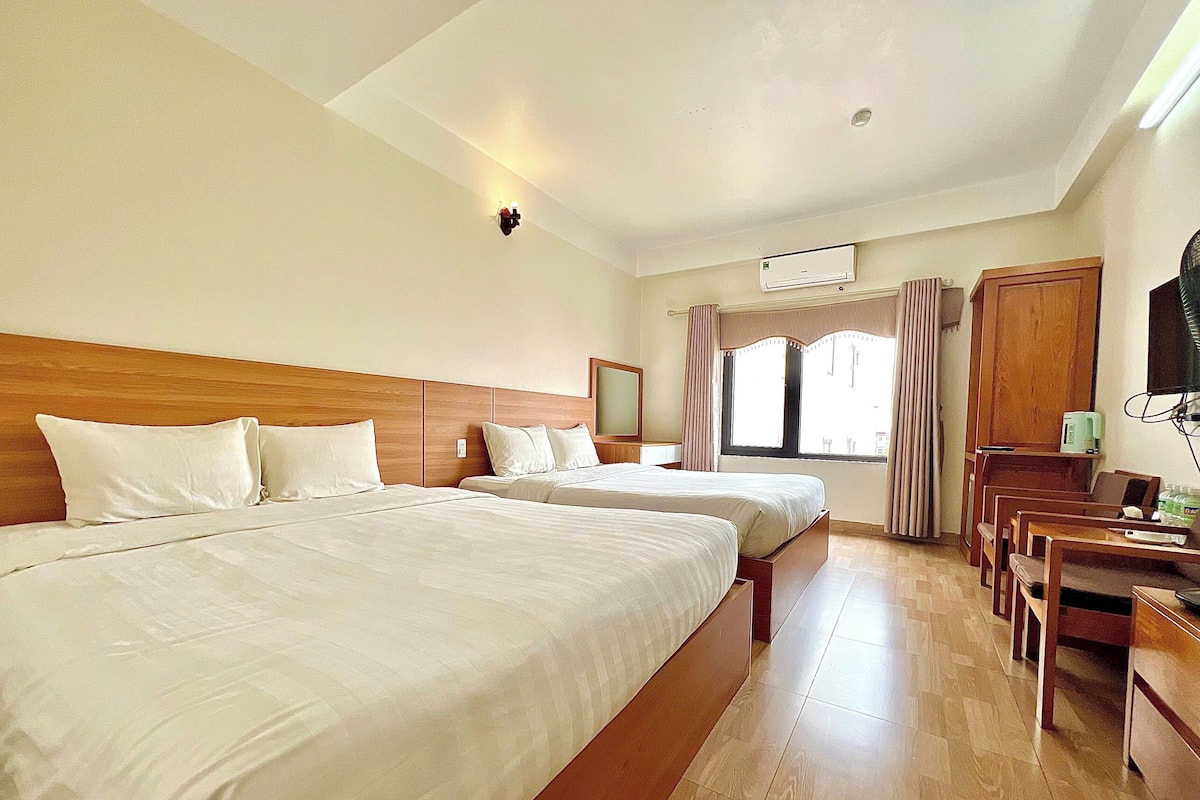 A hotel room with two beds, a wooden headboard, a desk with chairs, a flat-screen TV, and a window with curtains.