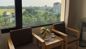 A room with a large window, a wooden table with chairs, a vase of flowers, and a standing fan.