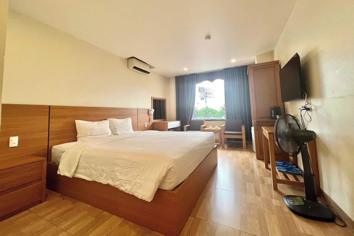 A hotel room with a large bed, a desk, a chair, a TV mounted on the wall, and a window with curtains.