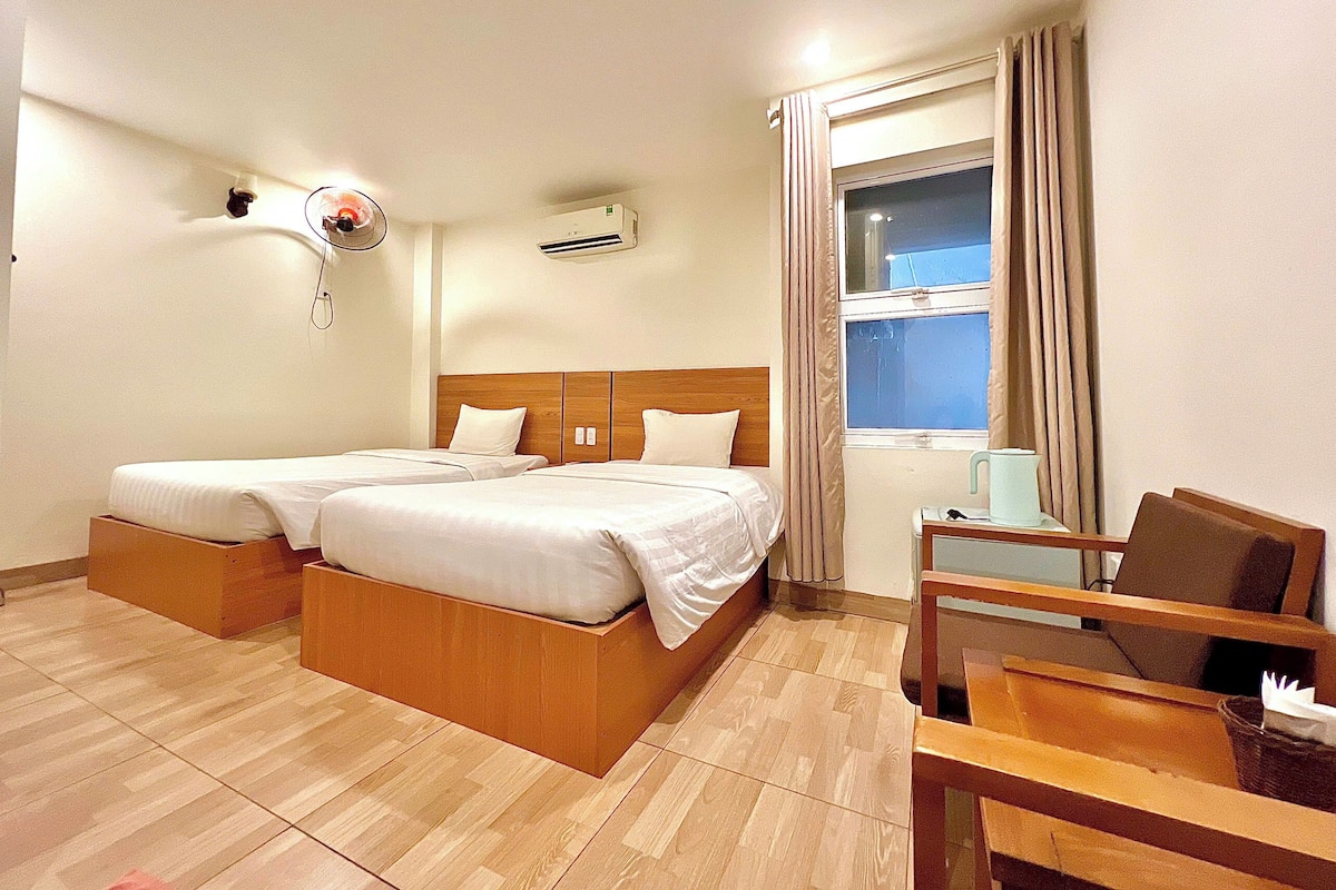 A hotel room with two beds, a wooden floor, a window with curtains, a fan, and a small table with a chair.