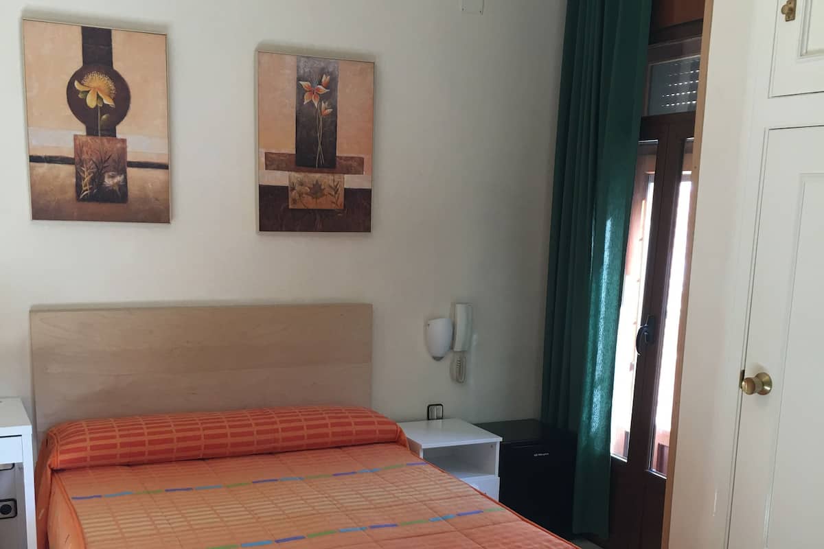 A neatly made bed with an orange patterned bedspread, a white bedside table with a lamp, and two wall-mounted paintings.