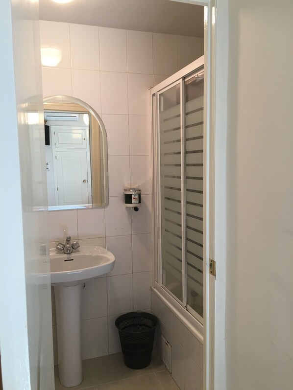 A small bathroom with a white sink, a mirror, a shower with frosted glass doors, and a black trash bin. - Hostal Cervantes (Valdemoro)
