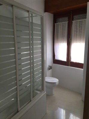 Un baño con inodoro, cabina de ducha de vidrio y ventana con cortinas.