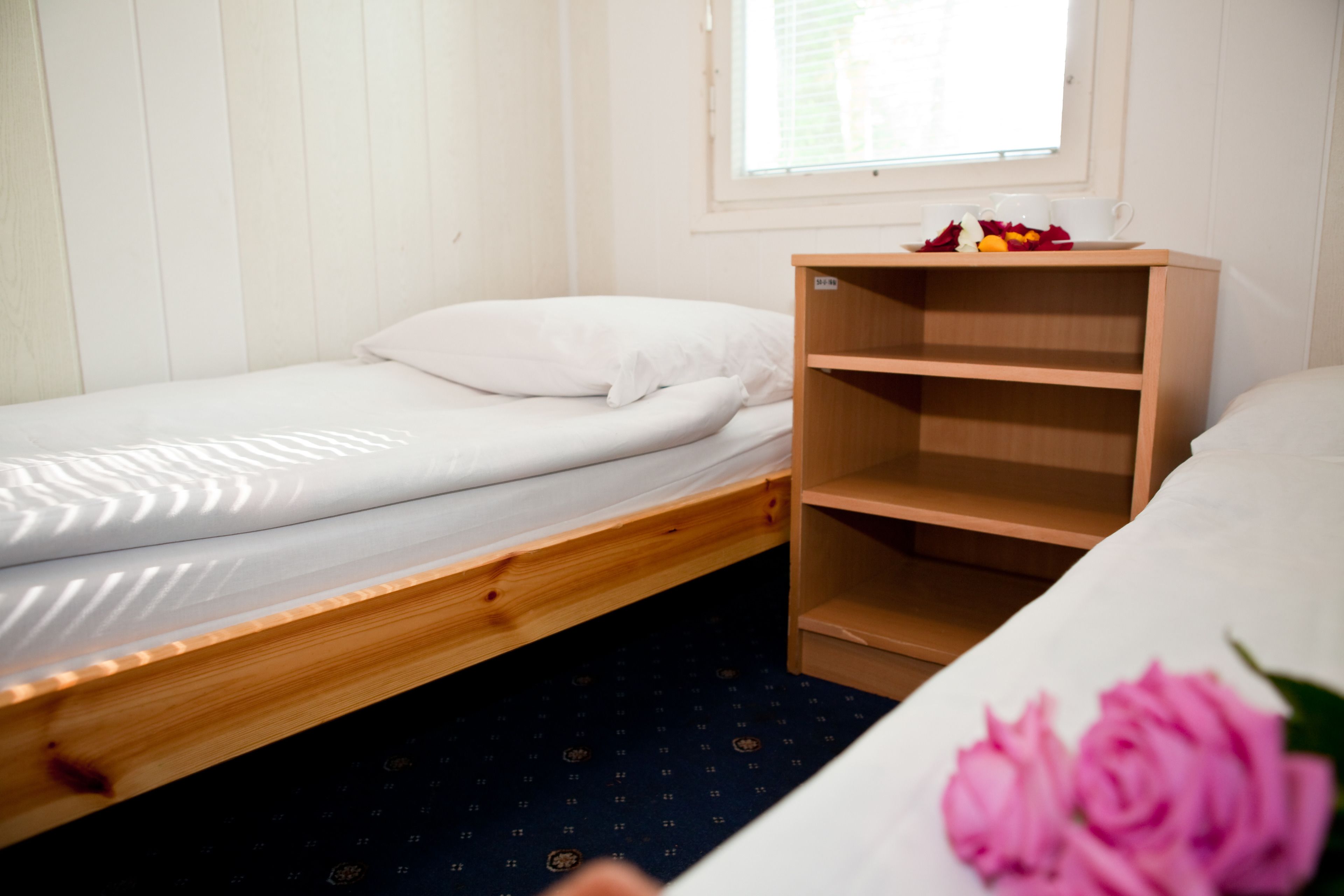 A single bed with a wooden frame, a white pillow, and a wooden nightstand with a cup and a plate of fruit.