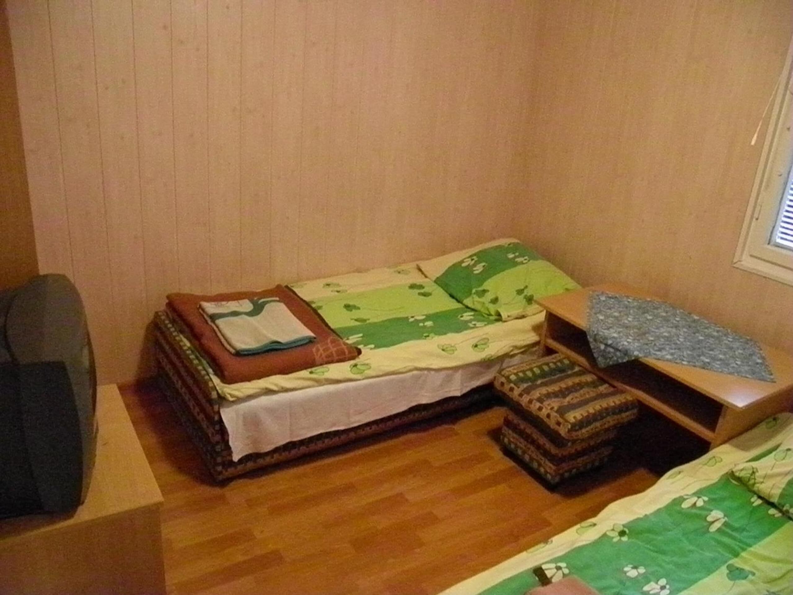 A small, single-bed room with a wooden floor, a bed with a green and white floral bedspread, a wooden nightstand, and a chair.