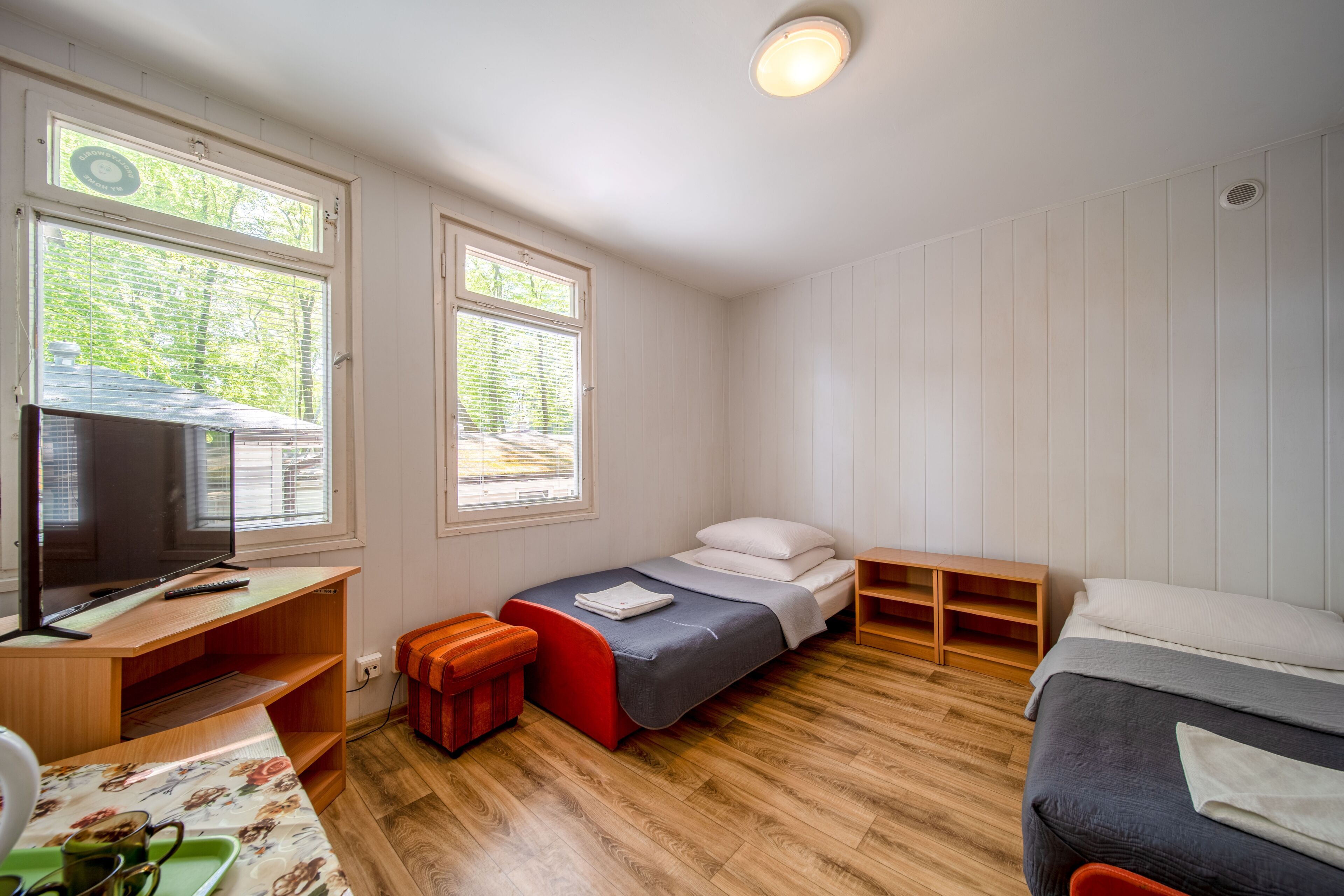 Quarto compacto com cama, escrivaninha e televisão, além de janela com persianas.