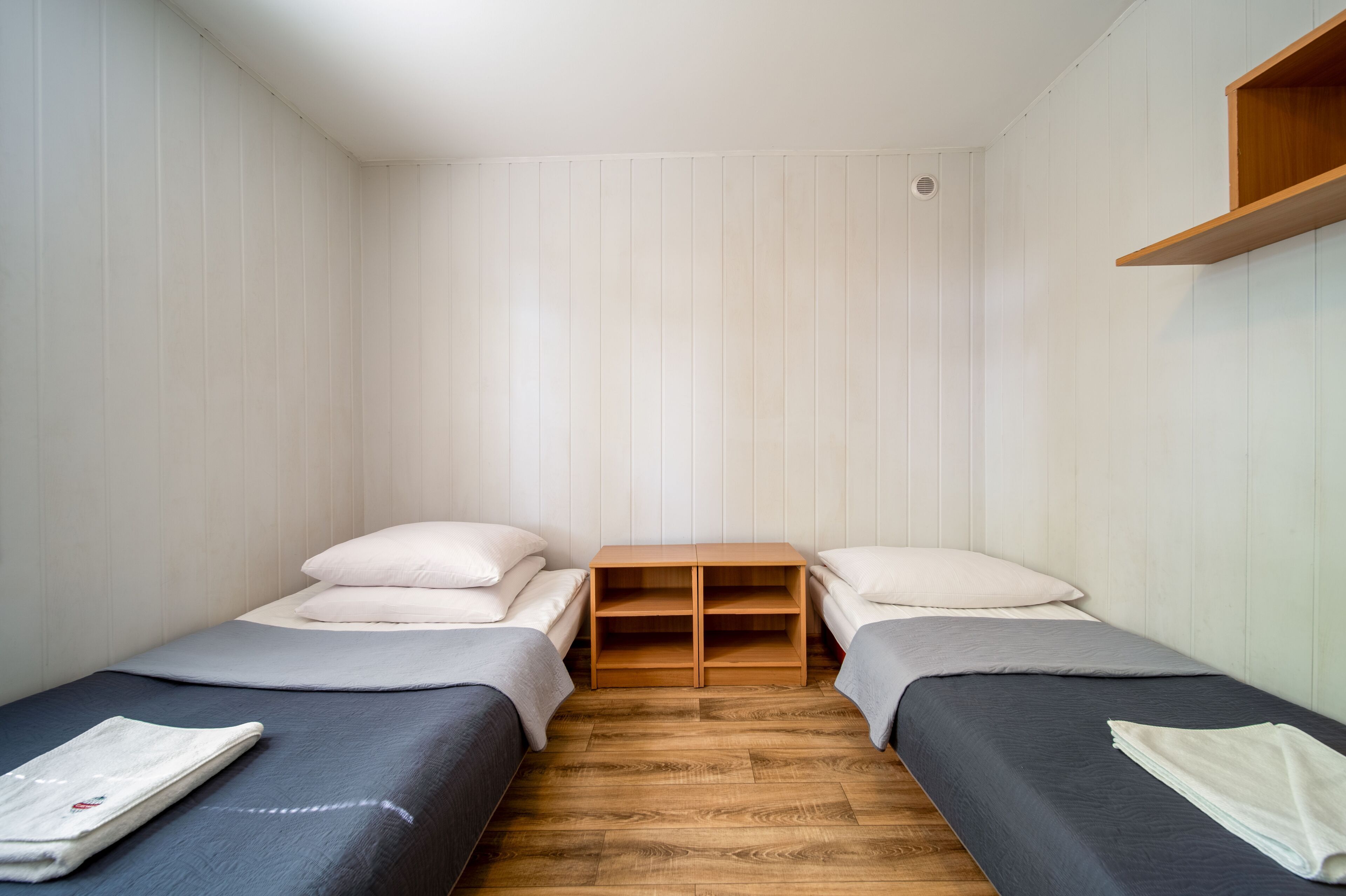 A small, clean room with two single beds, a wooden nightstand, and a white wall.