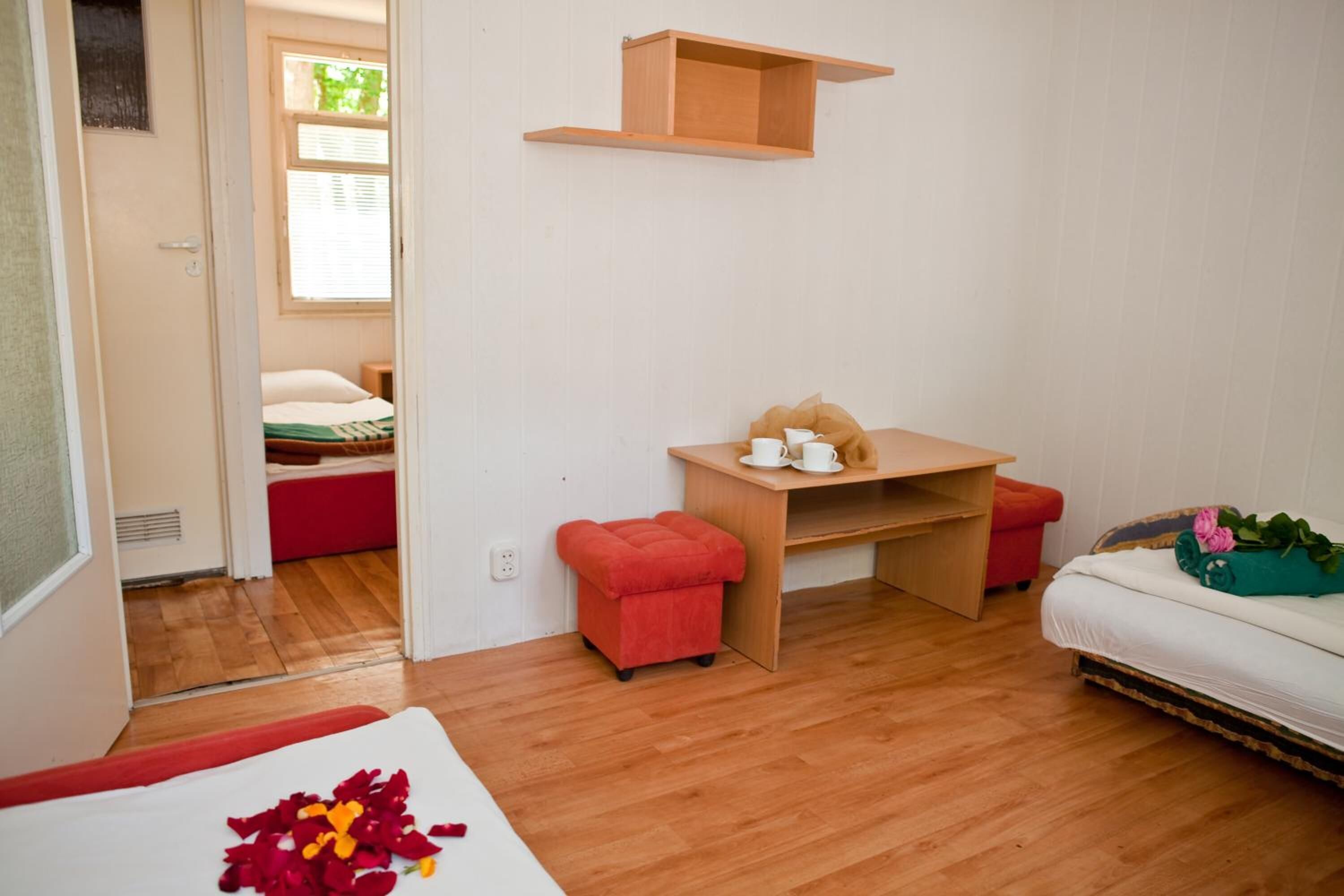 Een compacte kamer met een houten vloer, een klein bureau met stoel, een bed met een rode kussen en een raam met jaloezieën.