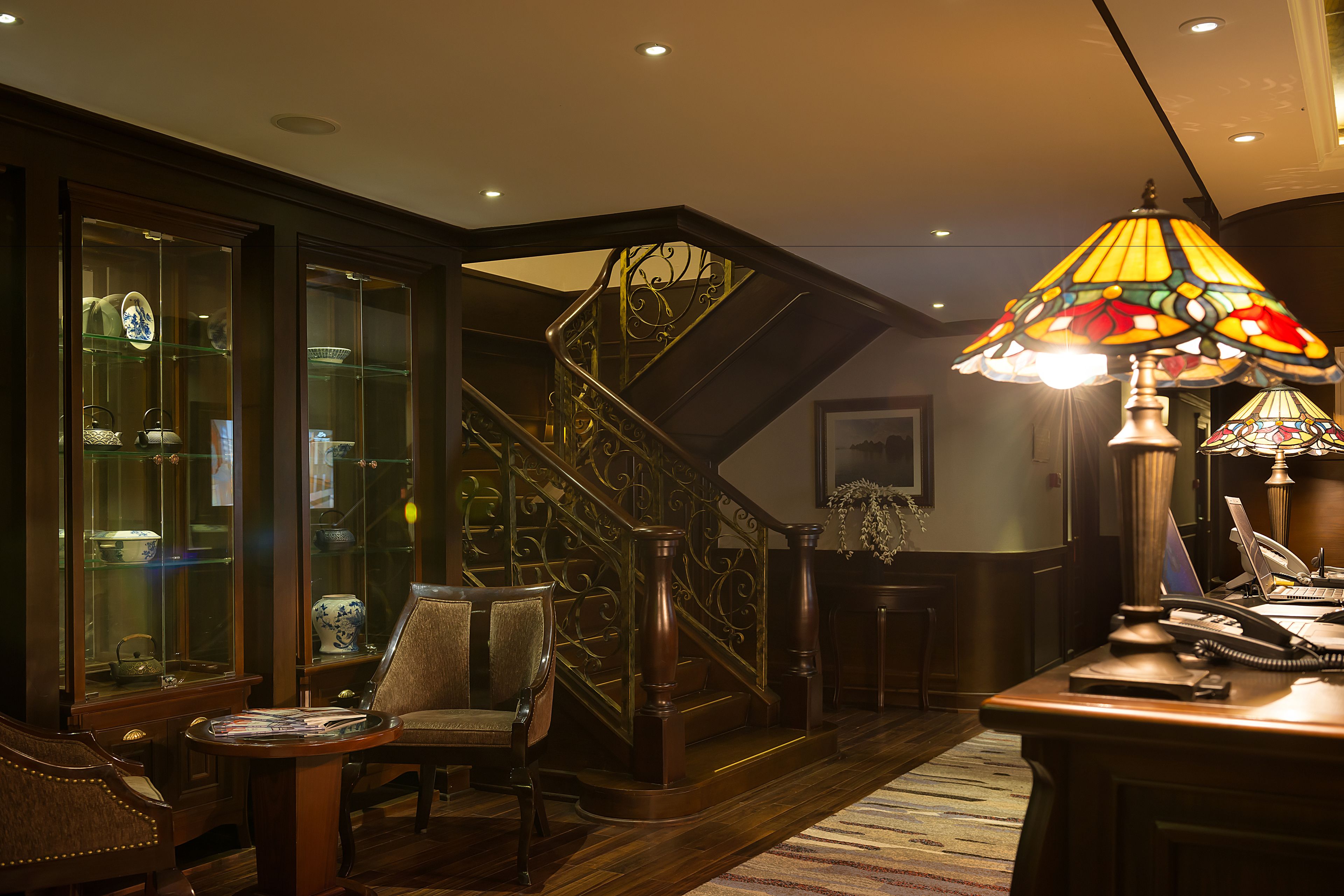 A staircase with ornate metal railings, a wooden desk with a lamp, and a chair in a warmly lit room with glass display cabinets.