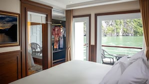 A hotel room with a large bed, a wardrobe, a window with a view of a body of water and cliffs, and a balcony with wicker furniture. - Paradise Elegance Cruise (Tuan Chau)