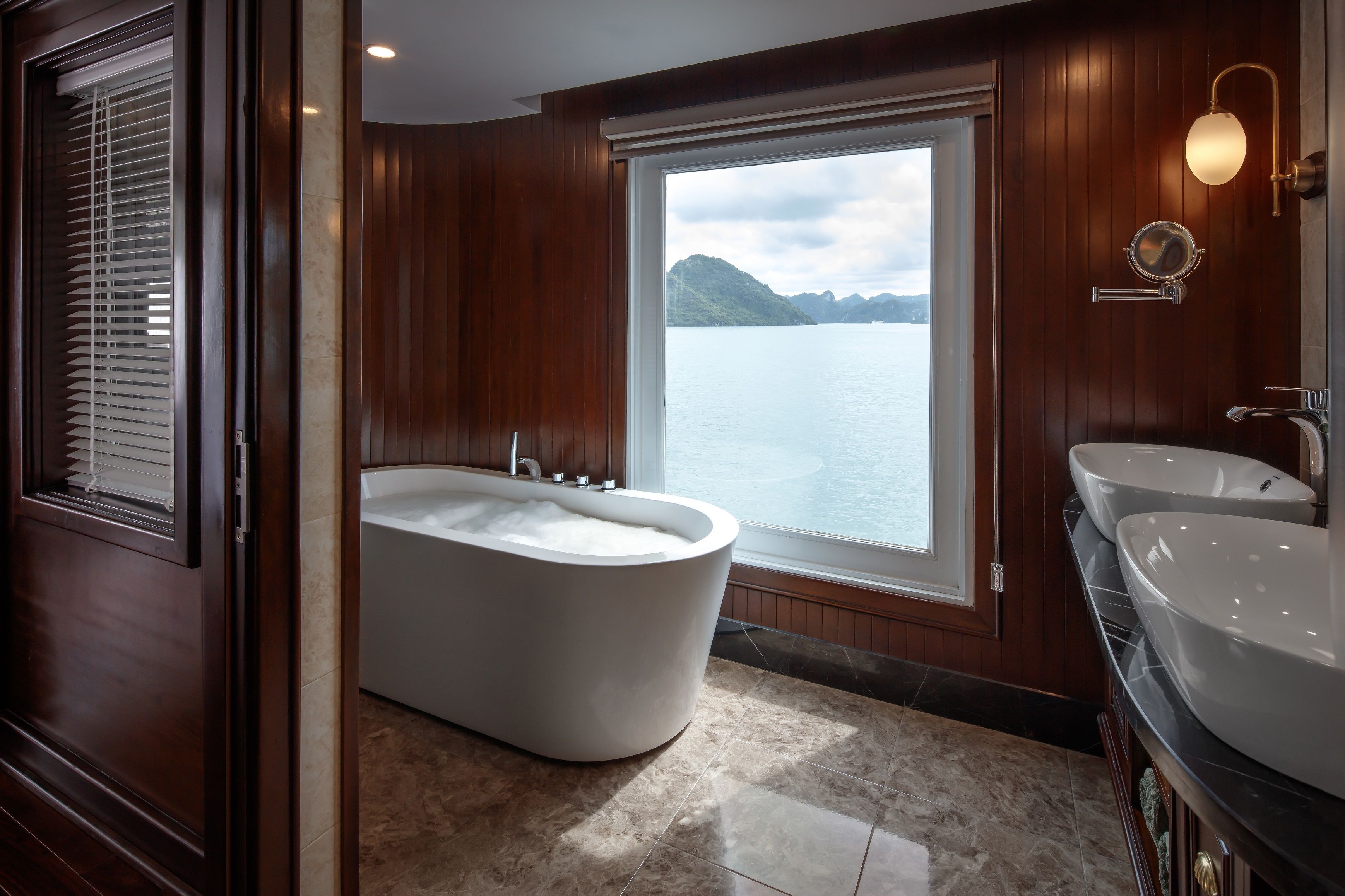 A modern bathroom with a freestanding bathtub, a double sink, and a large window offering a view of a lake and mountains.
