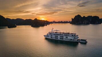 Un gran barco crucero fluvial con varias cubiertas y una bandera, rodeado de aguas tranquilas y formaciones rocosas distantes durante el atardecer.