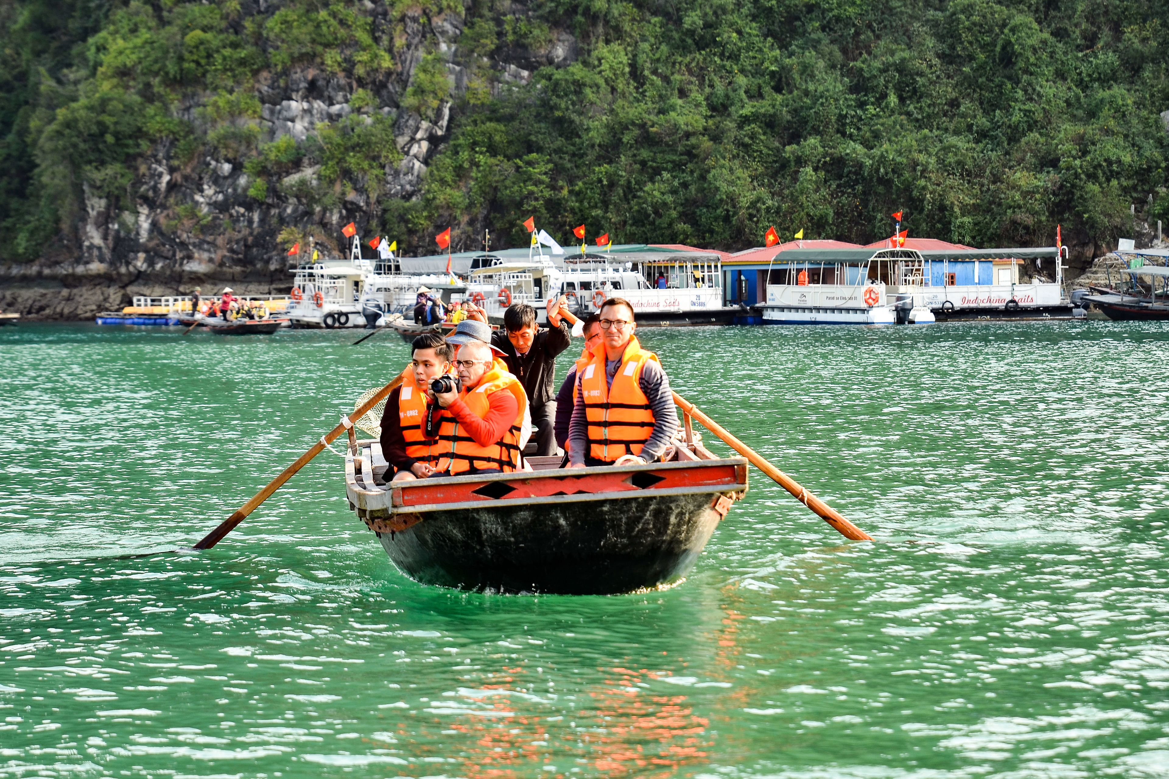 A boat with passengers wearing life jackets, paddling on a body of water with boats and a dock in the background.