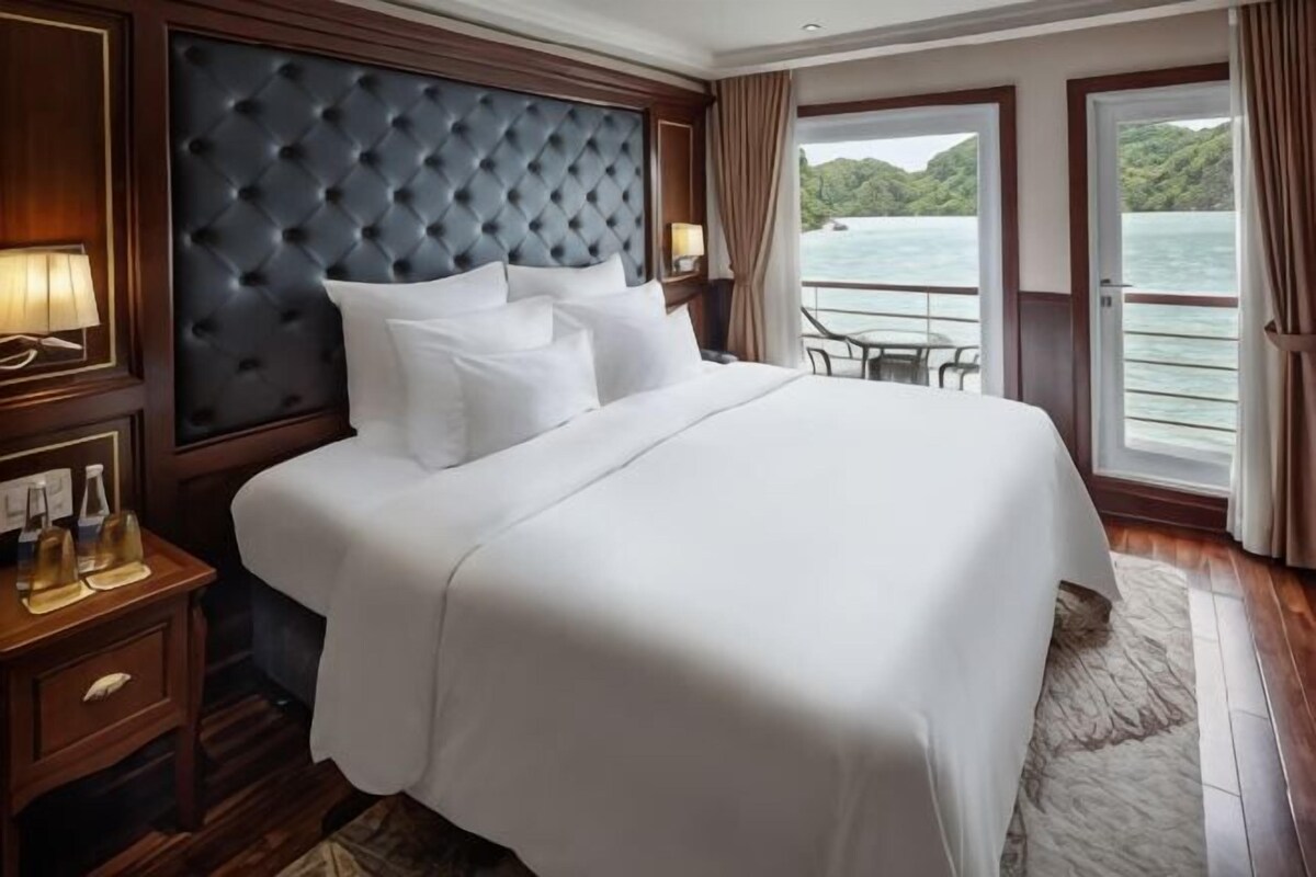 A large bed with a tufted headboard, a nightstand with a lamp, and a view of the water through a balcony door.