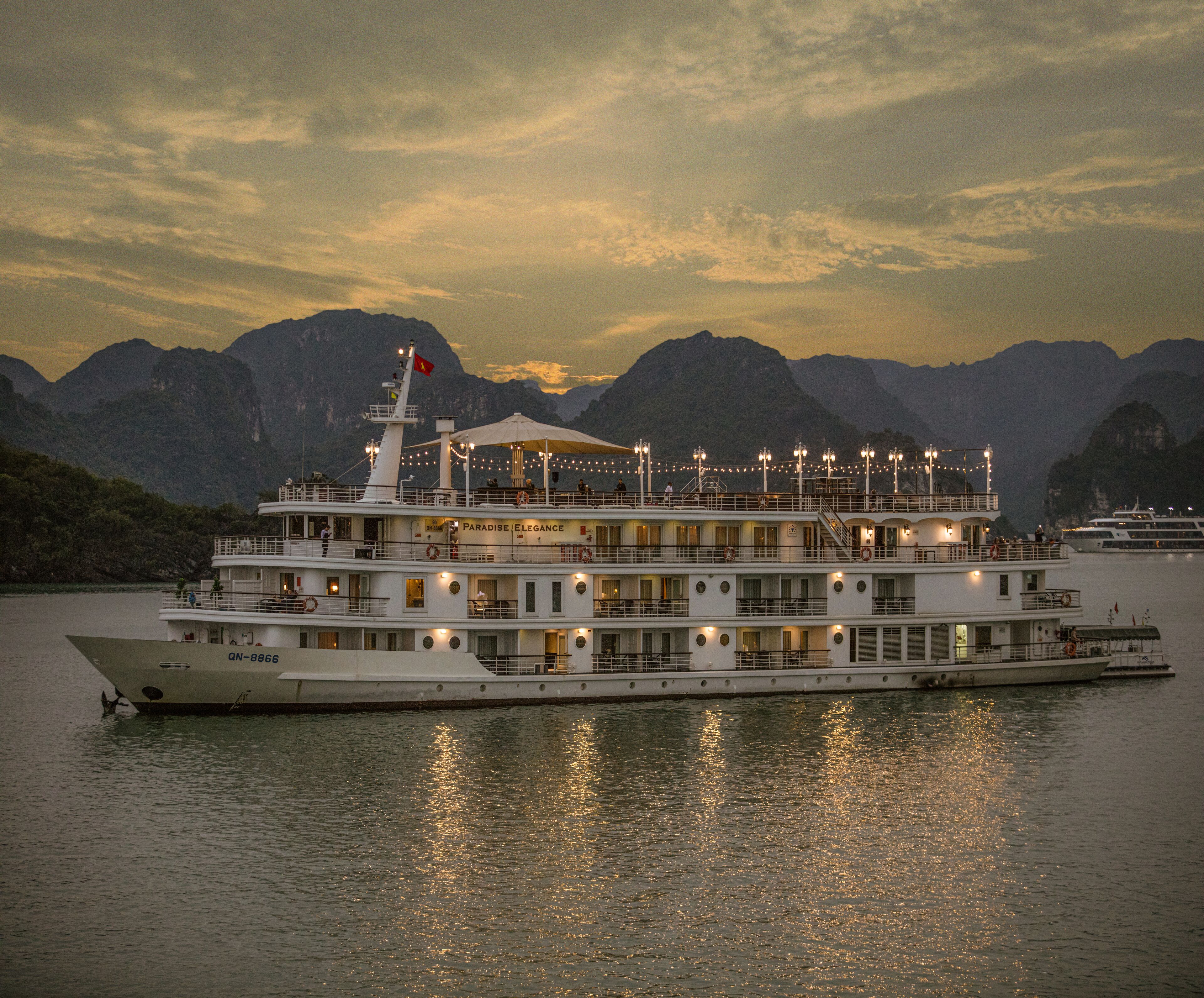 A large riverboat with multiple decks and a flag, docked at a harbor with mountains in the background.