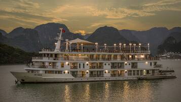 A large riverboat with multiple decks and a flag, docked at a harbor with mountains in the background.