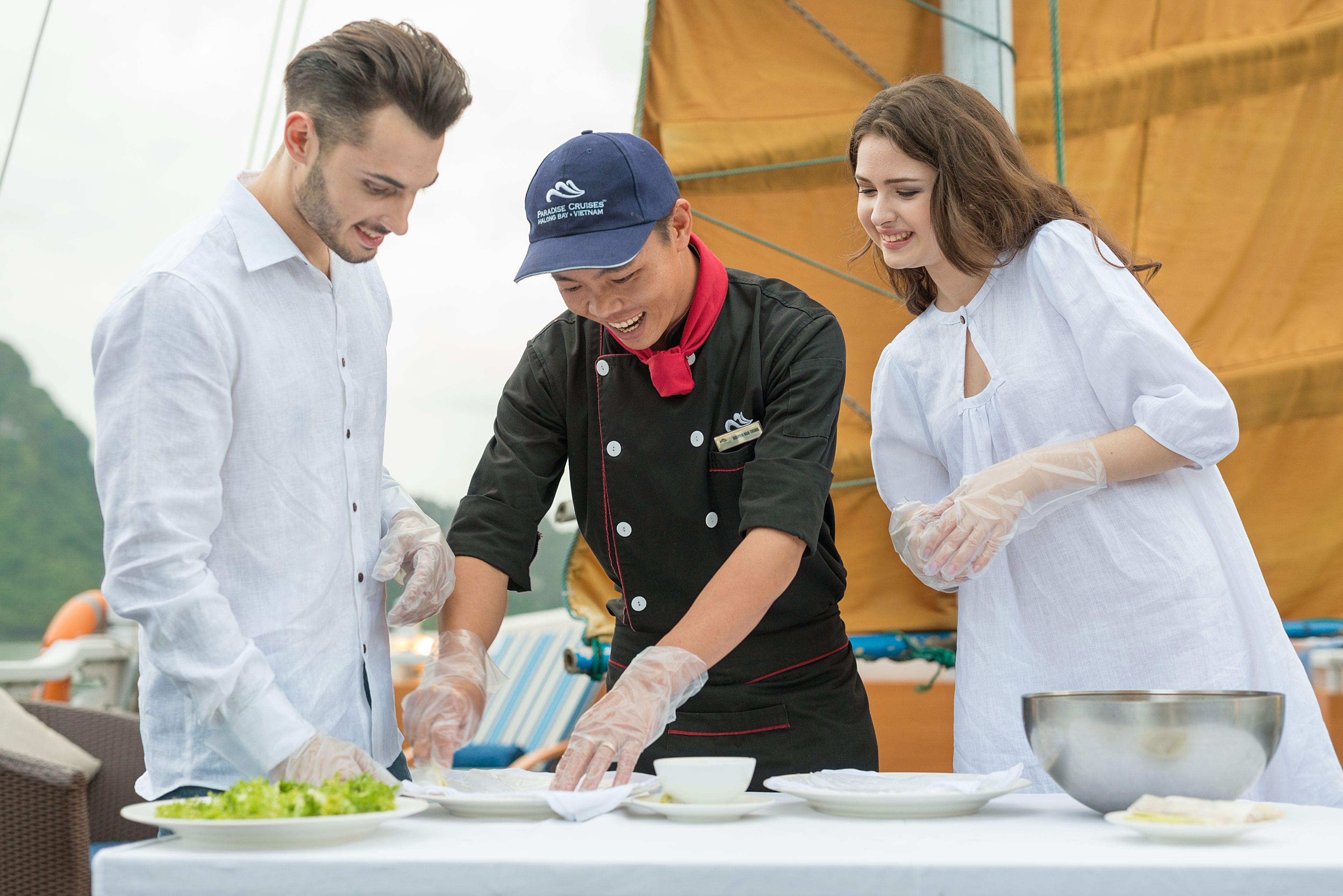 Un groupe de personnes sert des plats à une table en plein air, avec un voilier en arrière-plan.