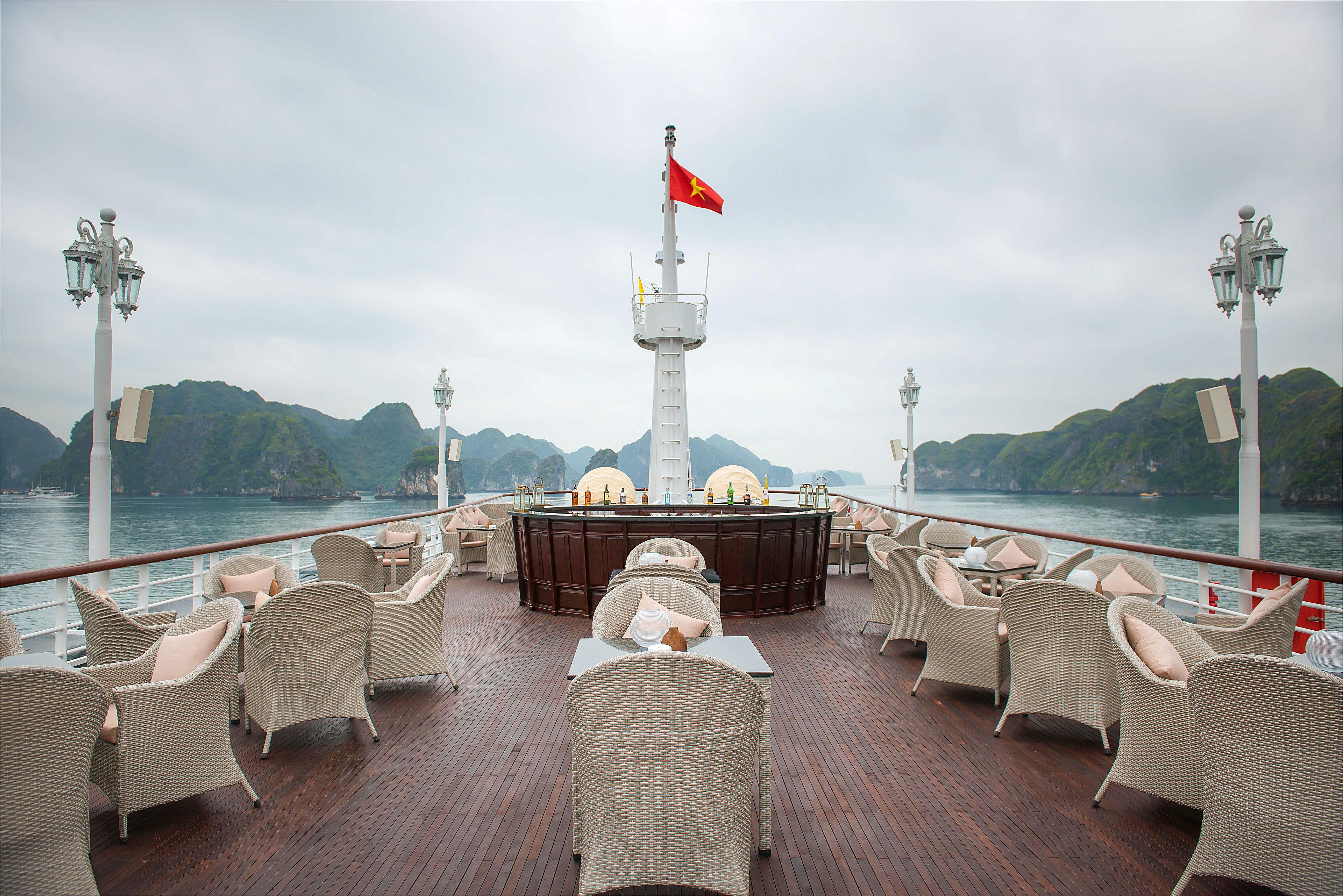 A deck with wicker furniture and a central bar area, overlooking a body of water with mountains in the background.