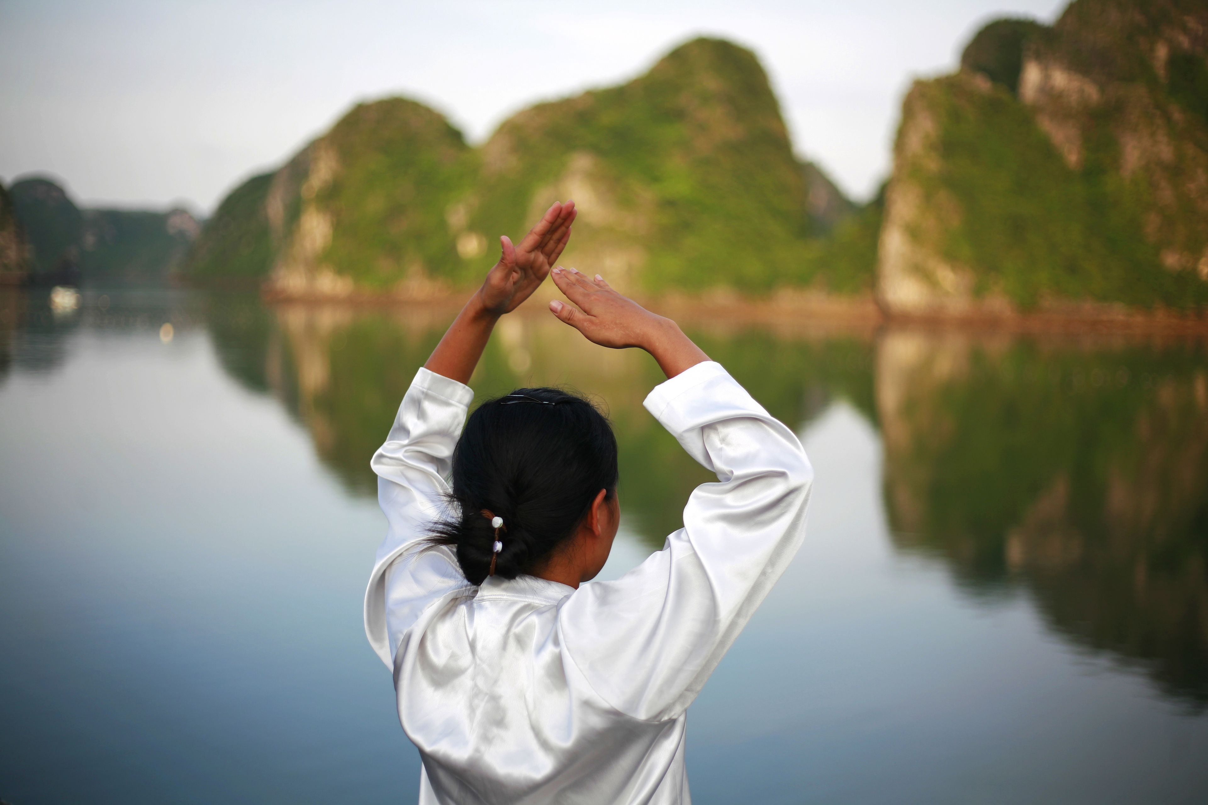 A person in a white shirt with their arms raised, standing by a calm body of water with green hills in the background.