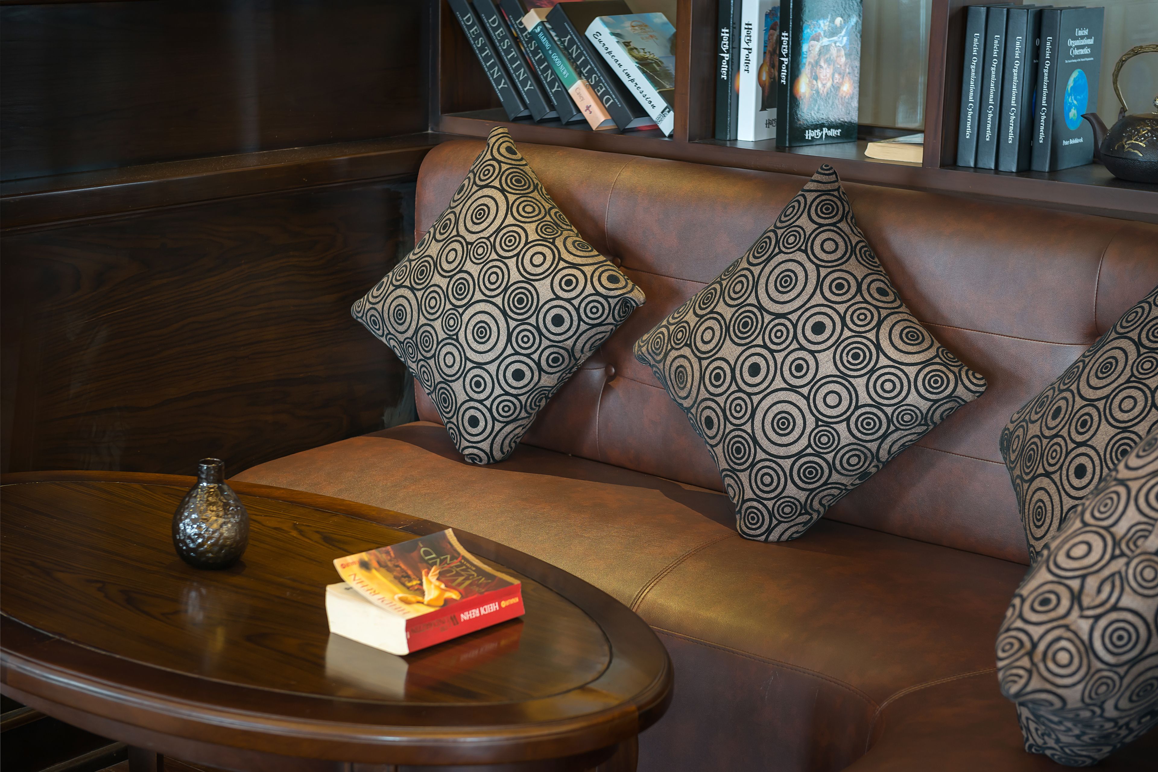 A leather sofa with patterned cushions, a small table with a book and a vase, and a bookshelf in the background.