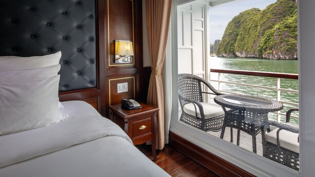 A hotel room with a bed, bedside table, lamp, and a view of a body of water and greenery. - Paradise Elegance Cruise (Tuan Chau)