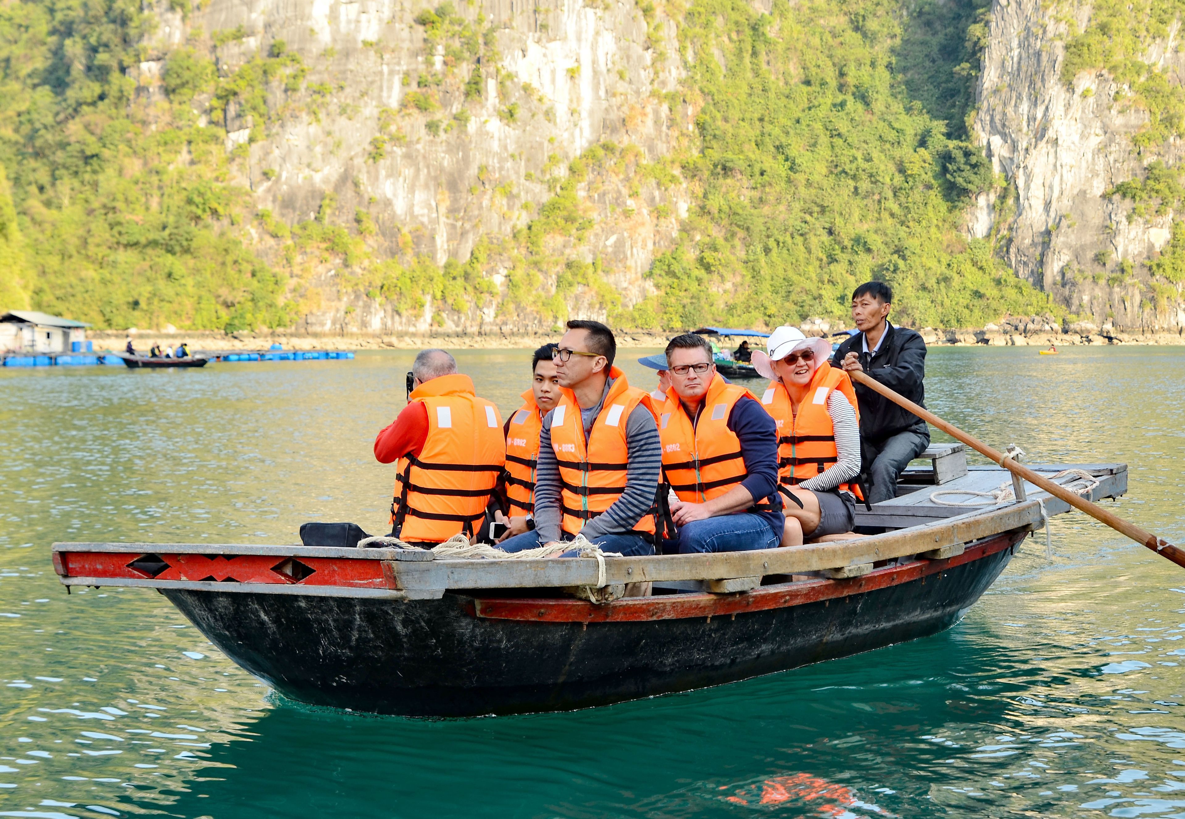 A boat with passengers wearing life jackets, a wooden oar, and a rocky cliff in the background.