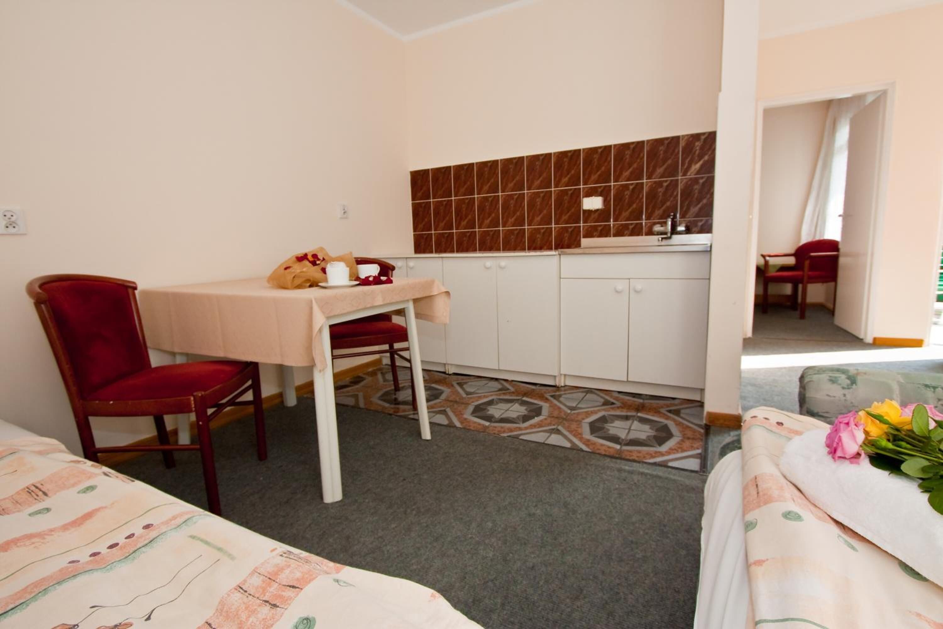 A small, cozy room with a dining table, a red chair, a kitchenette, and a bed with a floral bedspread.