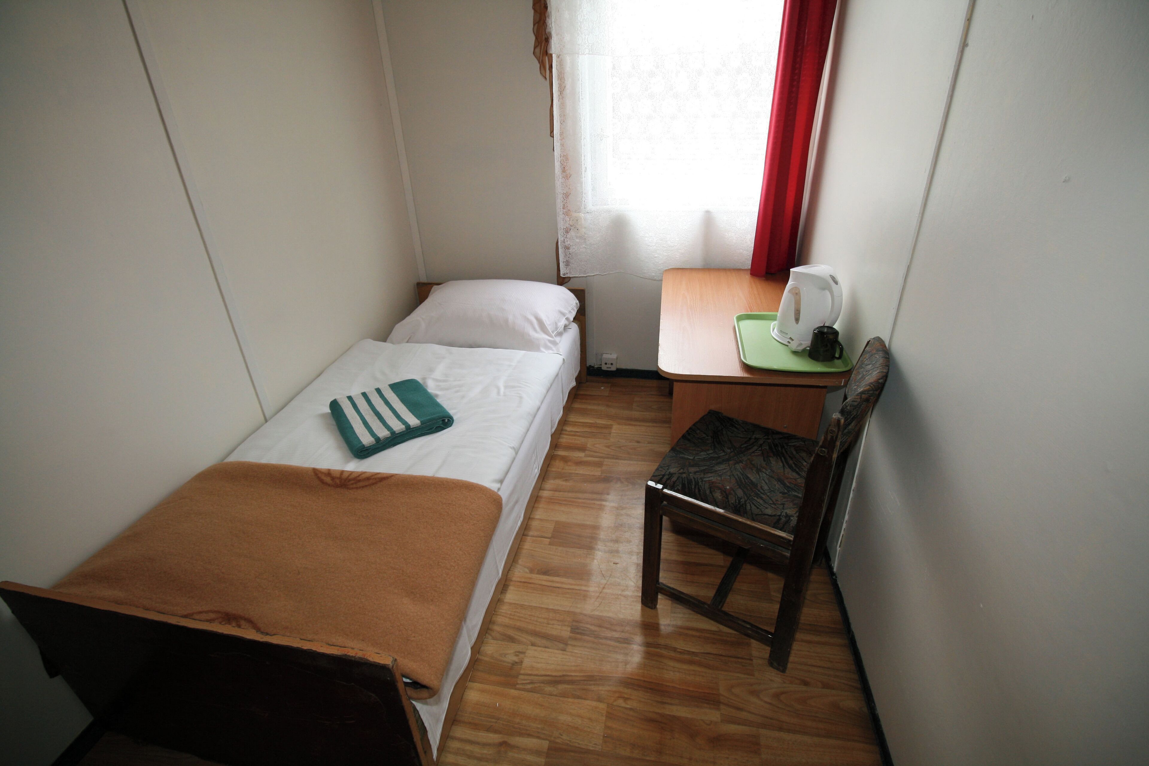 A small, single-bed room with a wooden floor, a desk with a chair, a kettle, and a window with curtains.