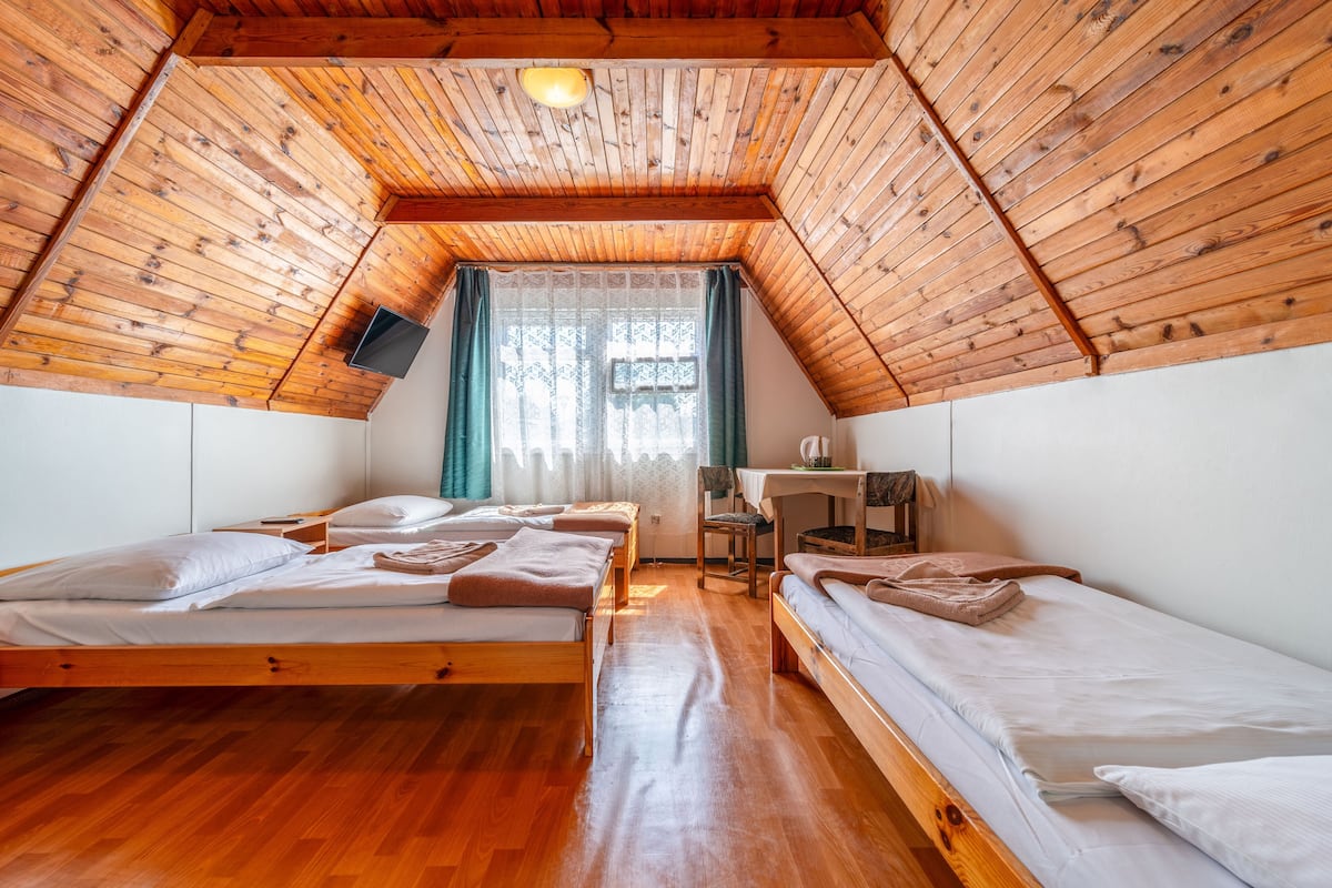 A room with wooden ceilings and floors, two beds, a desk, and a window with curtains.