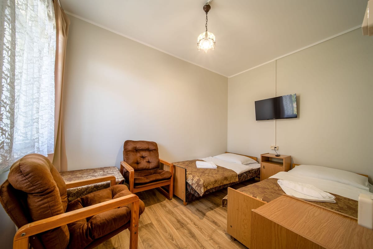 A hotel room with a bed, a chair, a small table, a TV mounted on the wall, and a window with curtains.