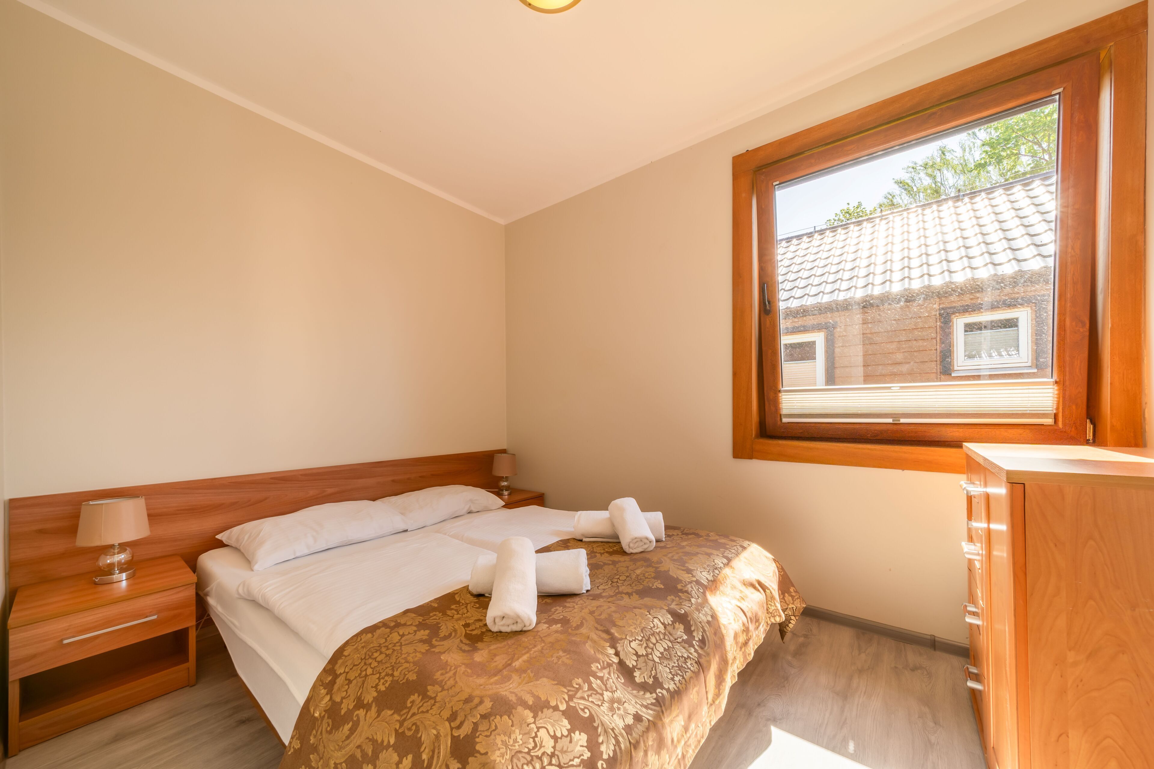 Une chambre avec une tête de lit en bois, un lit recouvert de draps blancs, deux serviettes enroulées, une table de chevet avec une lampe, une commode en bois et une fenêtre donnant sur un immeuble.
