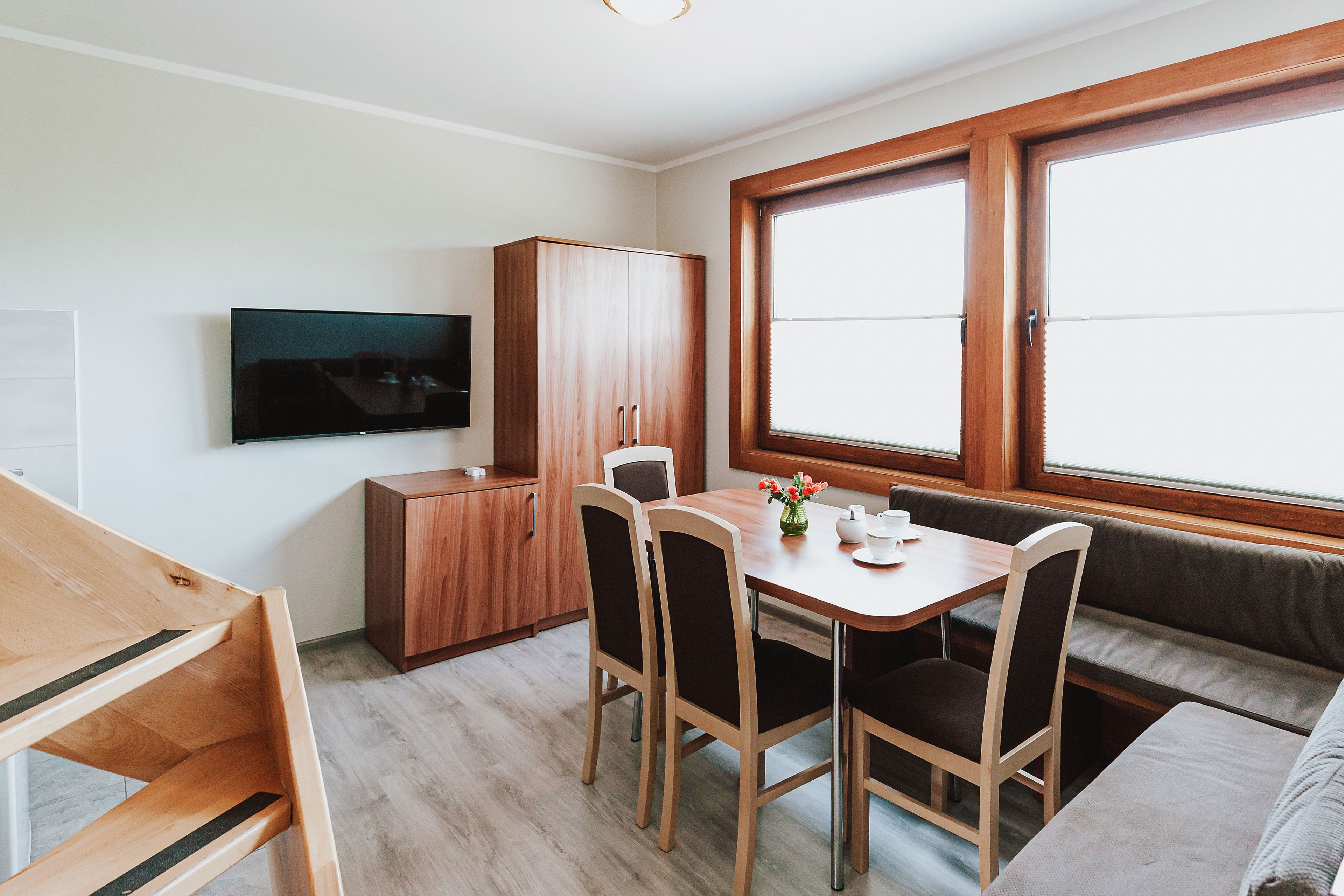 Una habitación compacta con una mesa de madera, cuatro sillas, un banco, un televisor montado en la pared y un ventanal grande.