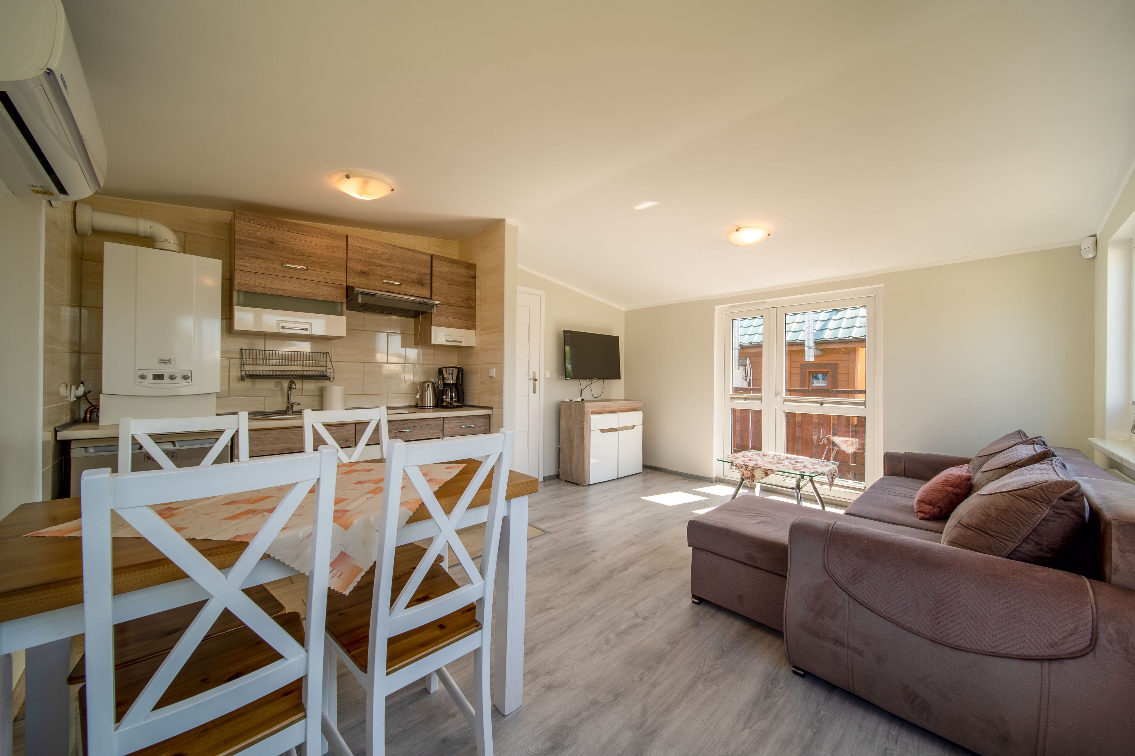 A modern kitchen with white cabinets, a dining area with a wooden table and white chairs, and a living area with a brown sofa and a TV.