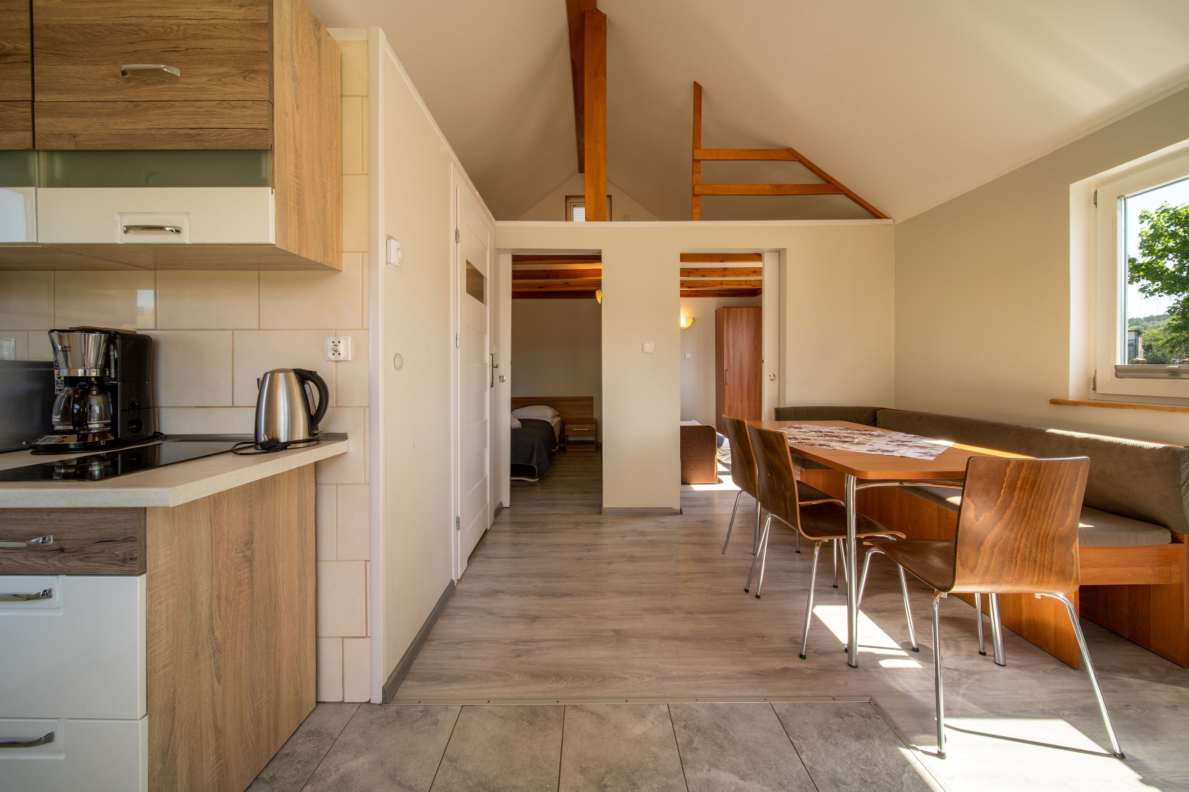 Eine moderne Küche mit Holzschränken, eine Essecke mit Holztisch und -stühlen und Blick in ein Schlafzimmer durch einen offenen Durchgang.