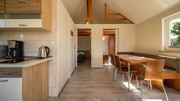 Eine moderne Küche mit Holzschränken, eine Essecke mit Holztisch und -stühlen und Blick in ein Schlafzimmer durch einen offenen Durchgang.