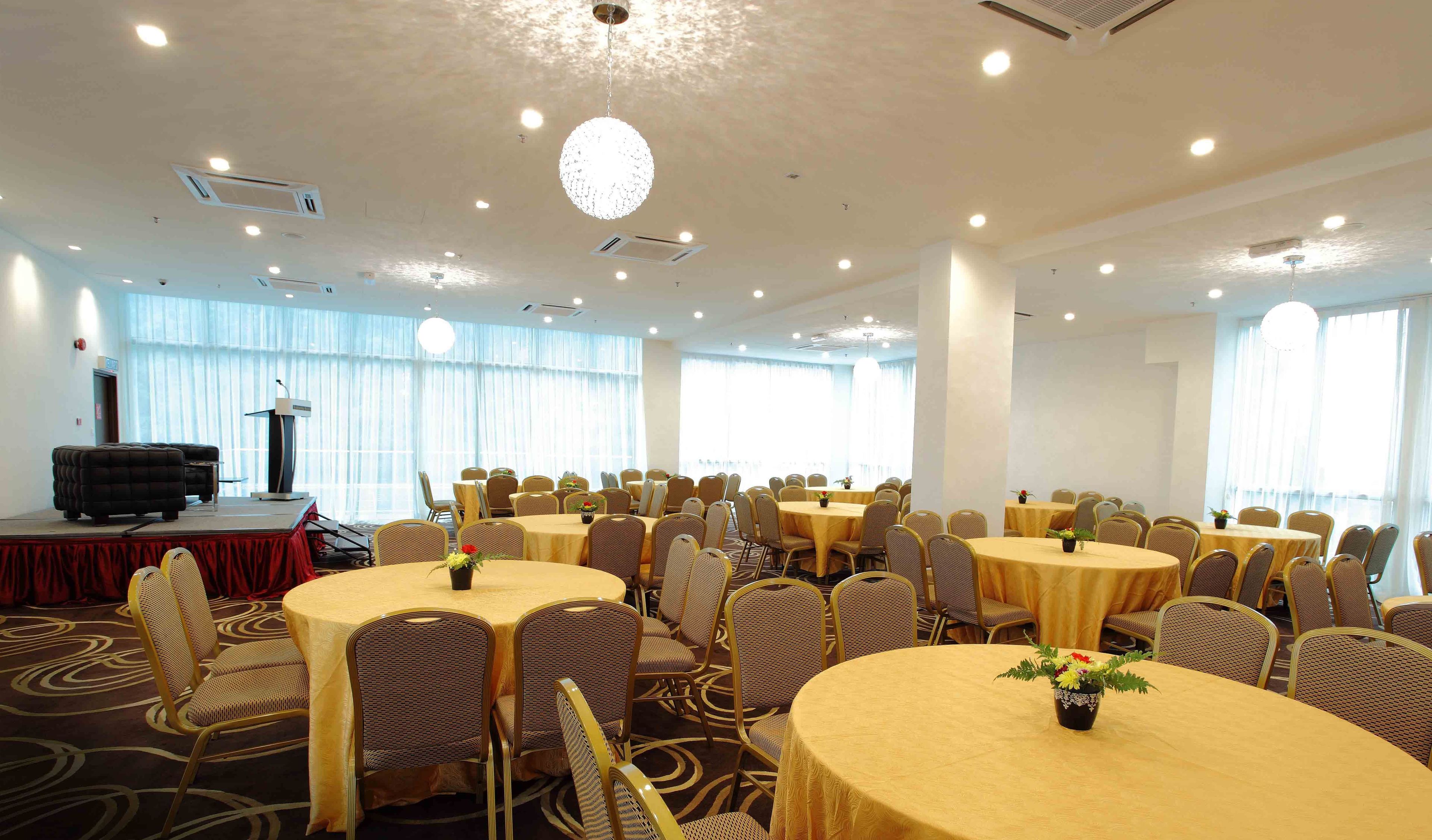 A conference room with round tables and chairs, a stage with a podium, and large windows with curtains.