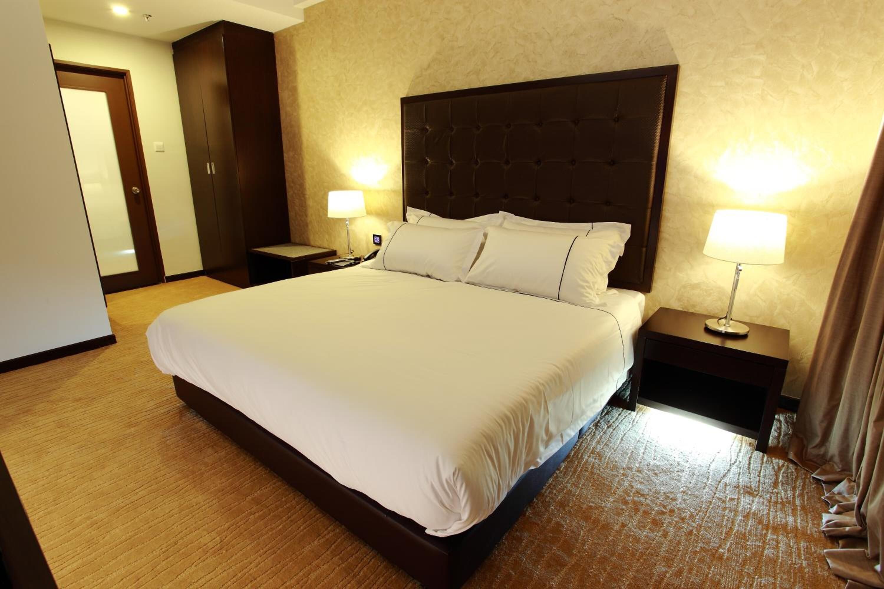 A hotel room with a large bed, two bedside tables with lamps, a wardrobe, and a door.