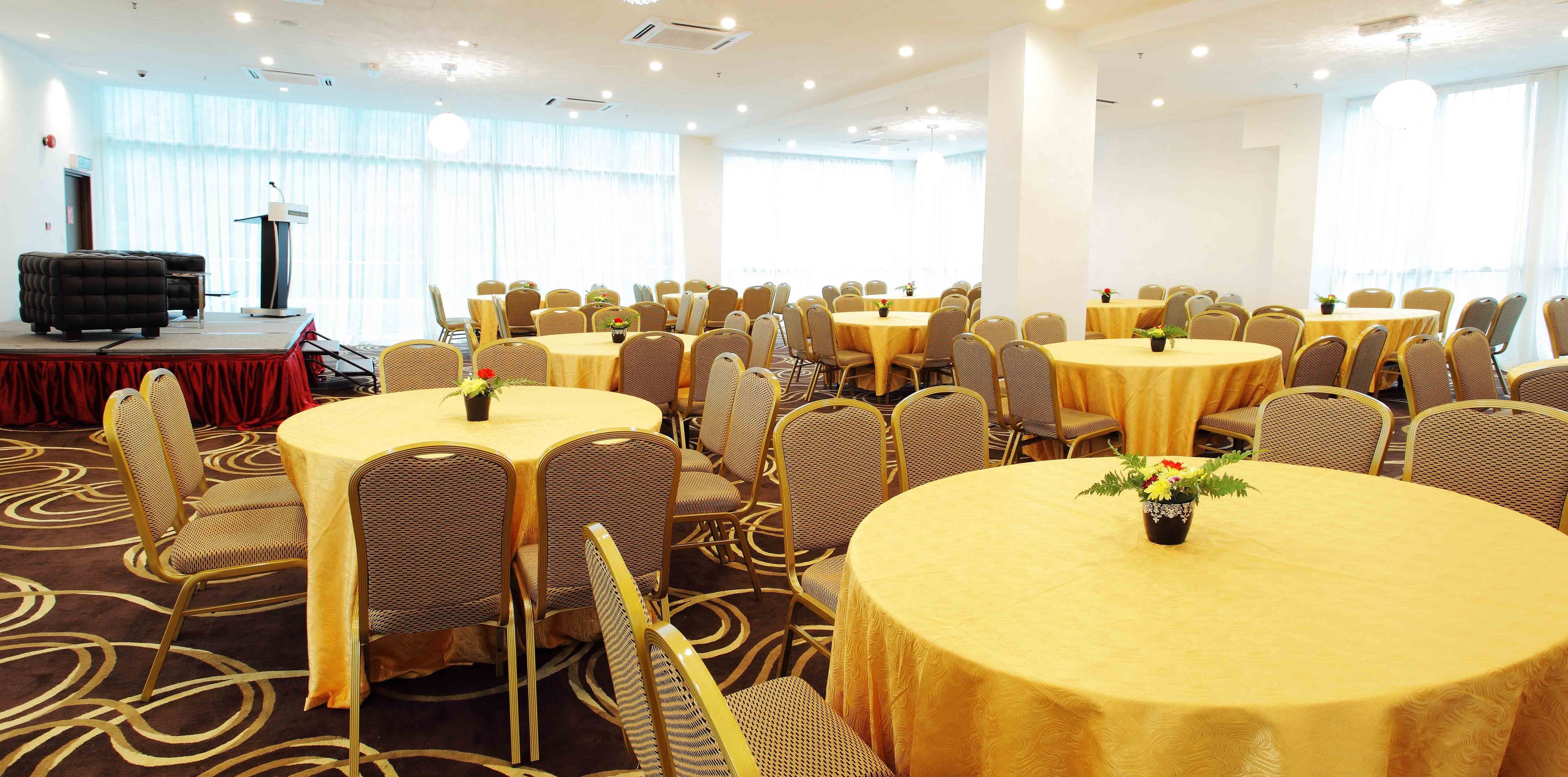 A conference room with multiple round tables and chairs, a stage with a podium, and large windows allowing natural light.