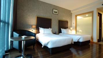 A hotel room with two beds, a small round table, a chair, and a large window with curtains.