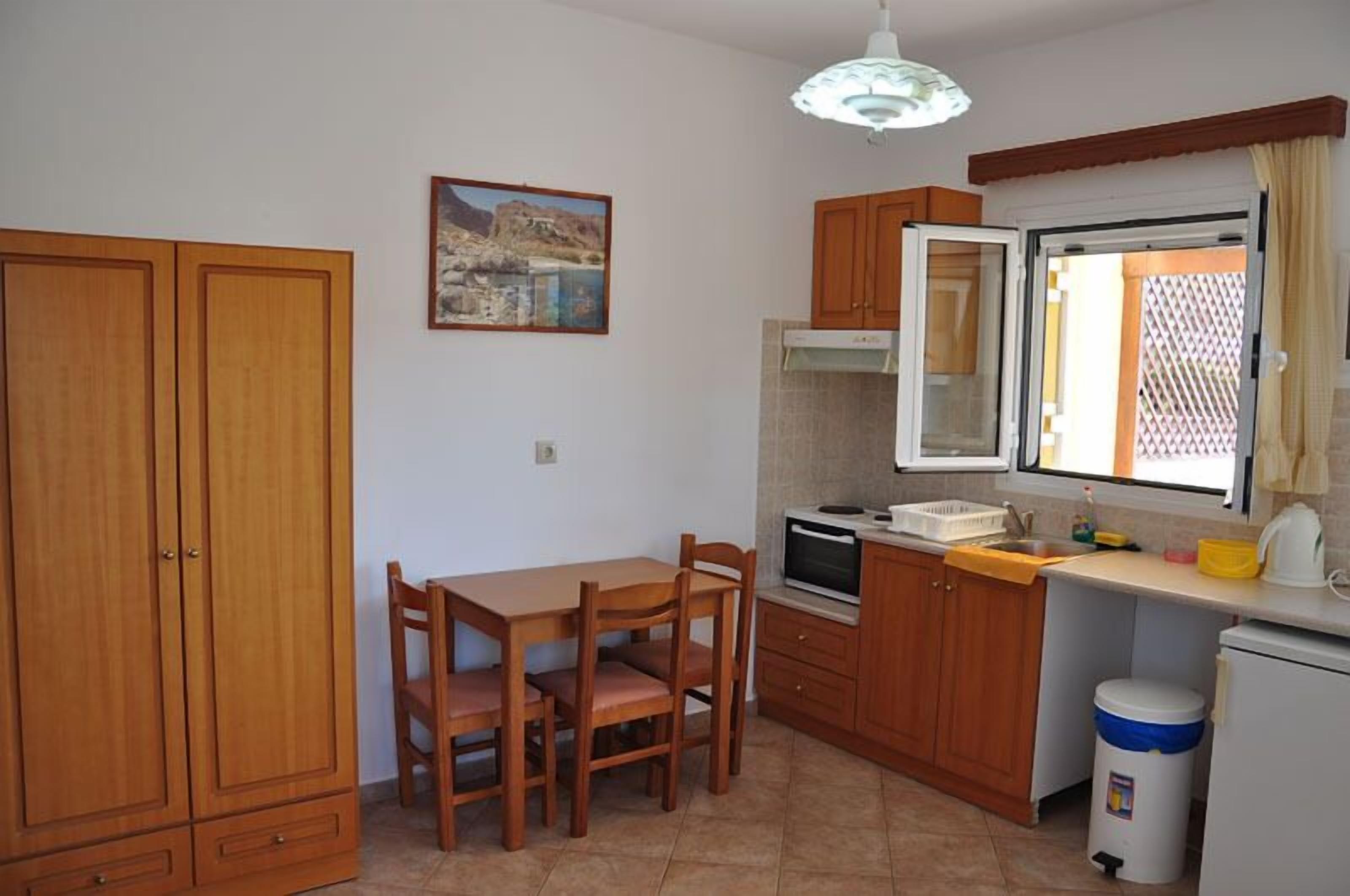 A compact kitchen with wooden cabinets, a dining table with chairs, a built-in oven, and a window with a view of a building.