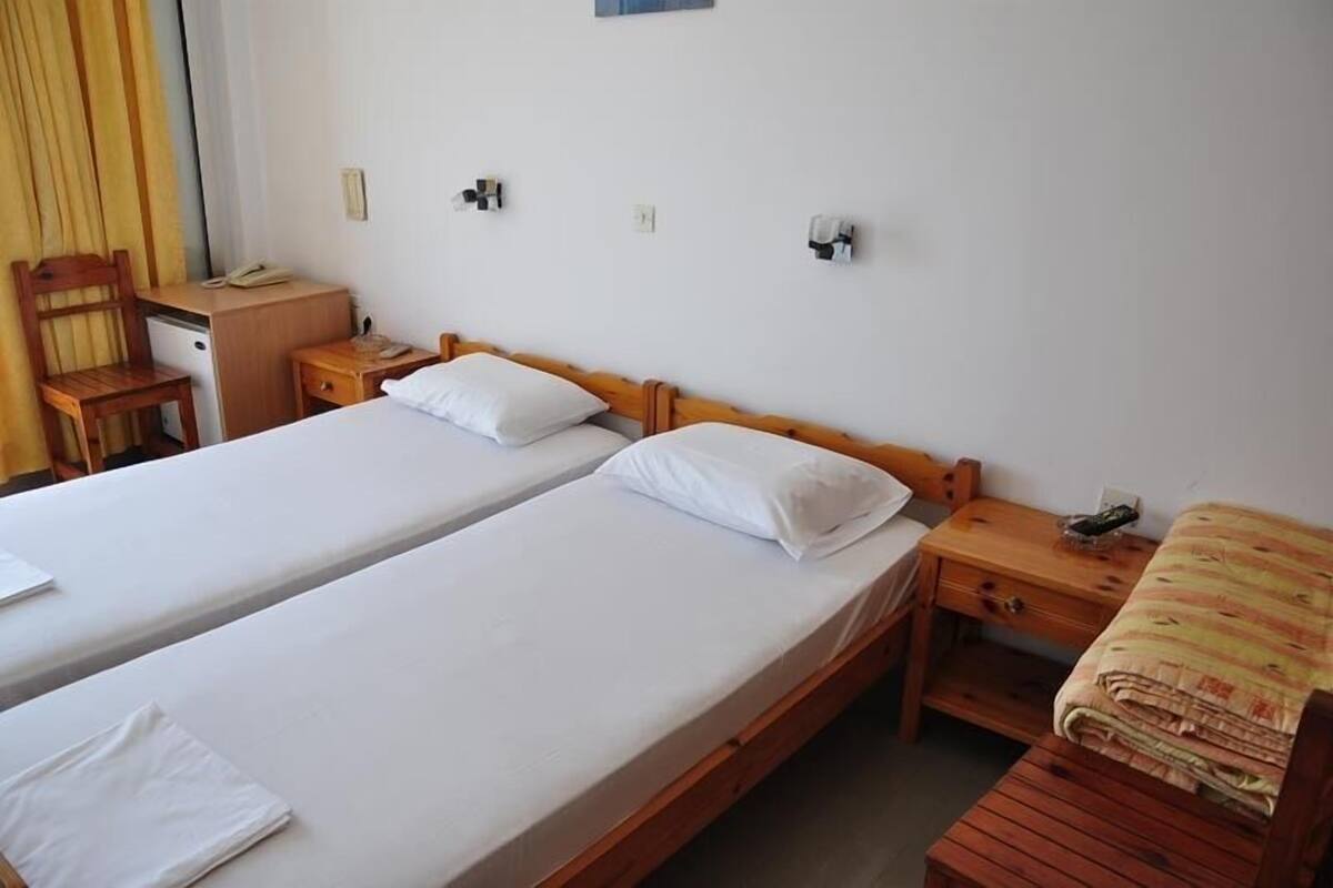 A hotel room with two single beds, a wooden desk, a chair, a small table, and a bedside table with a lamp.