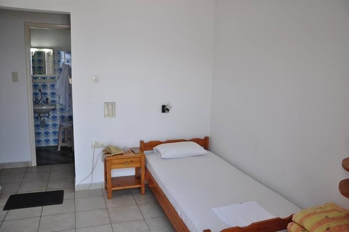 A small, clean room with a single bed, a wooden nightstand, and a small table. A bathroom is visible in the background with blue tiles.