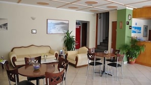 A hotel lobby with a beige sofa, wooden chairs, a round table with a vase, and a staircase leading to another room. - Nora Hotel (Sitia)